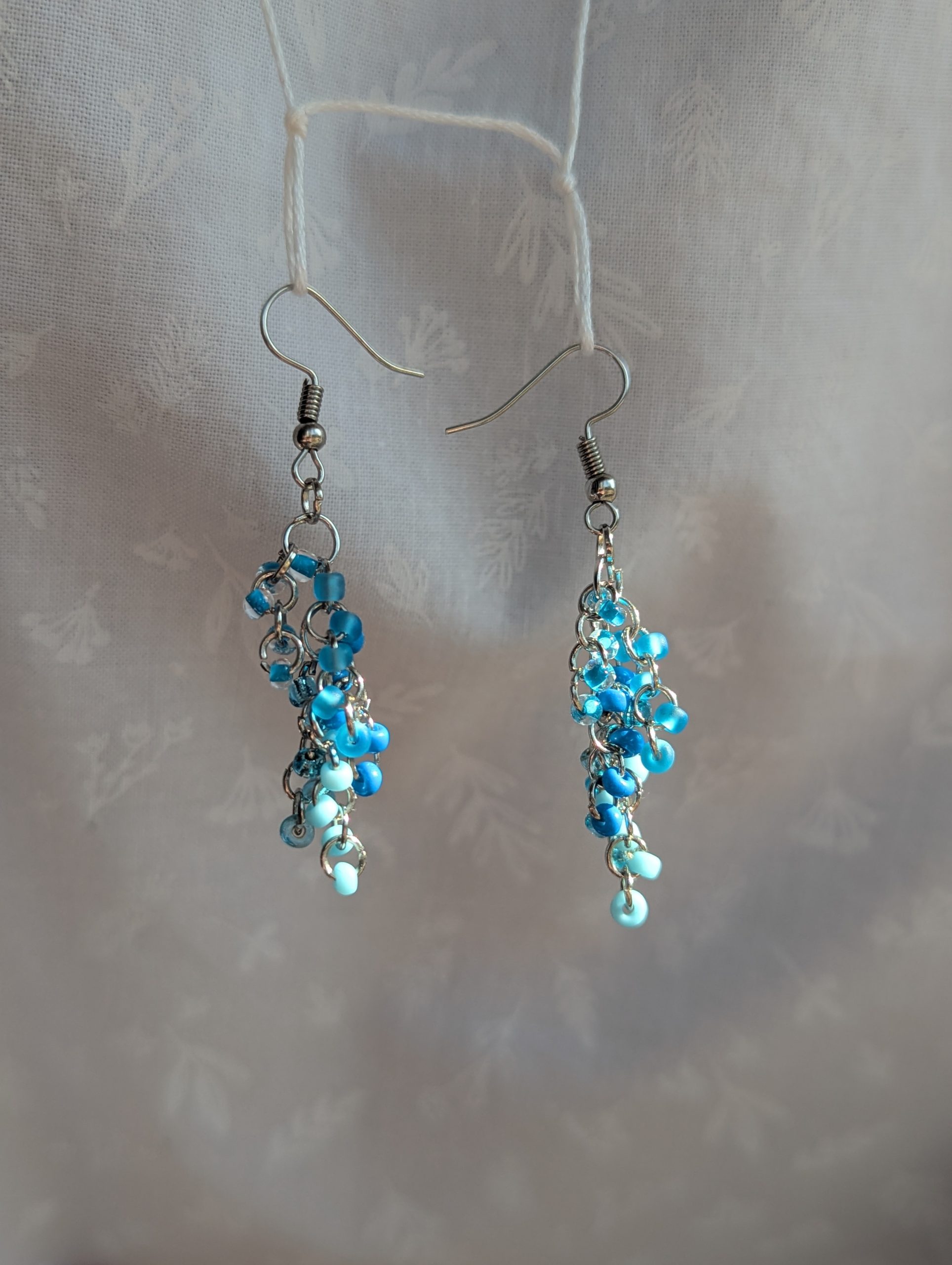 Blue beaded earrings with six different shades of blue glass seed beads, steel-colored jump rings, and steel earring hooks.