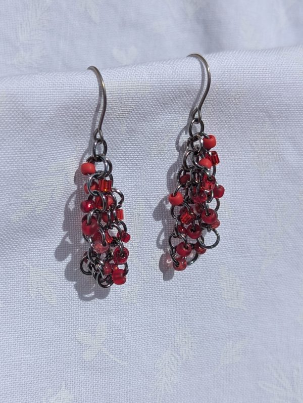 A pair of handmade red beaded dangle earrings with four shades of red glass seed beads attached to bronze-colored jump rings, hanging from titanium hooks.