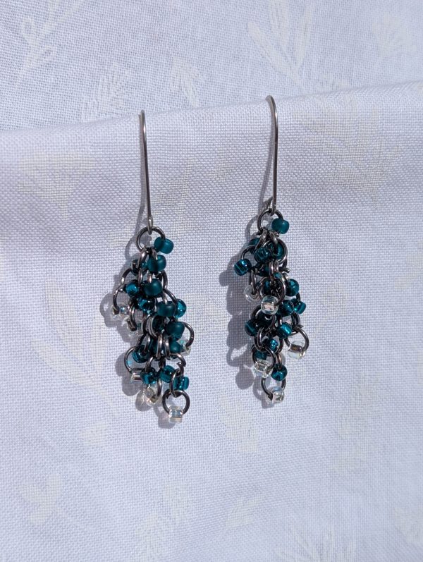 A pair of handmade dangle earrings with deep teal and clear glass seed beads attached to dark gunmetal-toned chains, hanging from silver-tone ear hooks.
