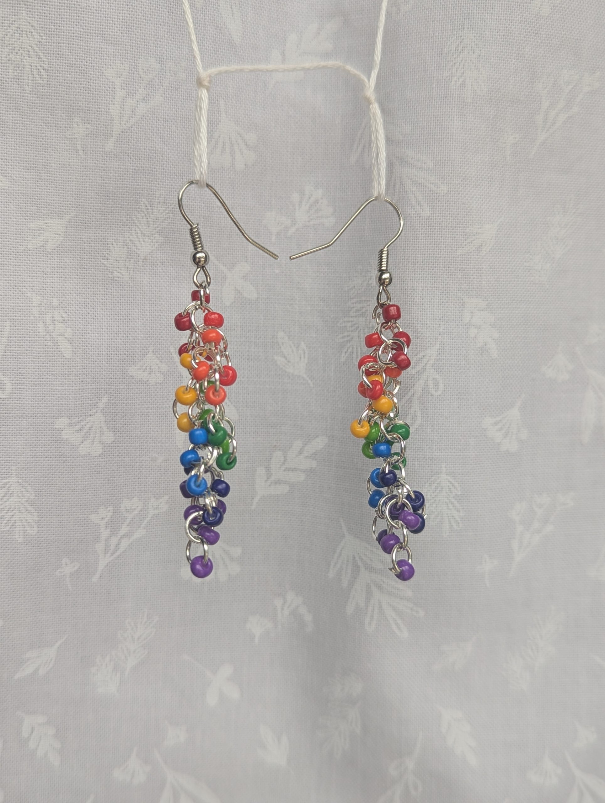 A pair of handmade rainbow dangle earrings featuring red, orange, yellow, green, blue, indigo, and purple glass seed beads with steel-colored hooks and jump rings.