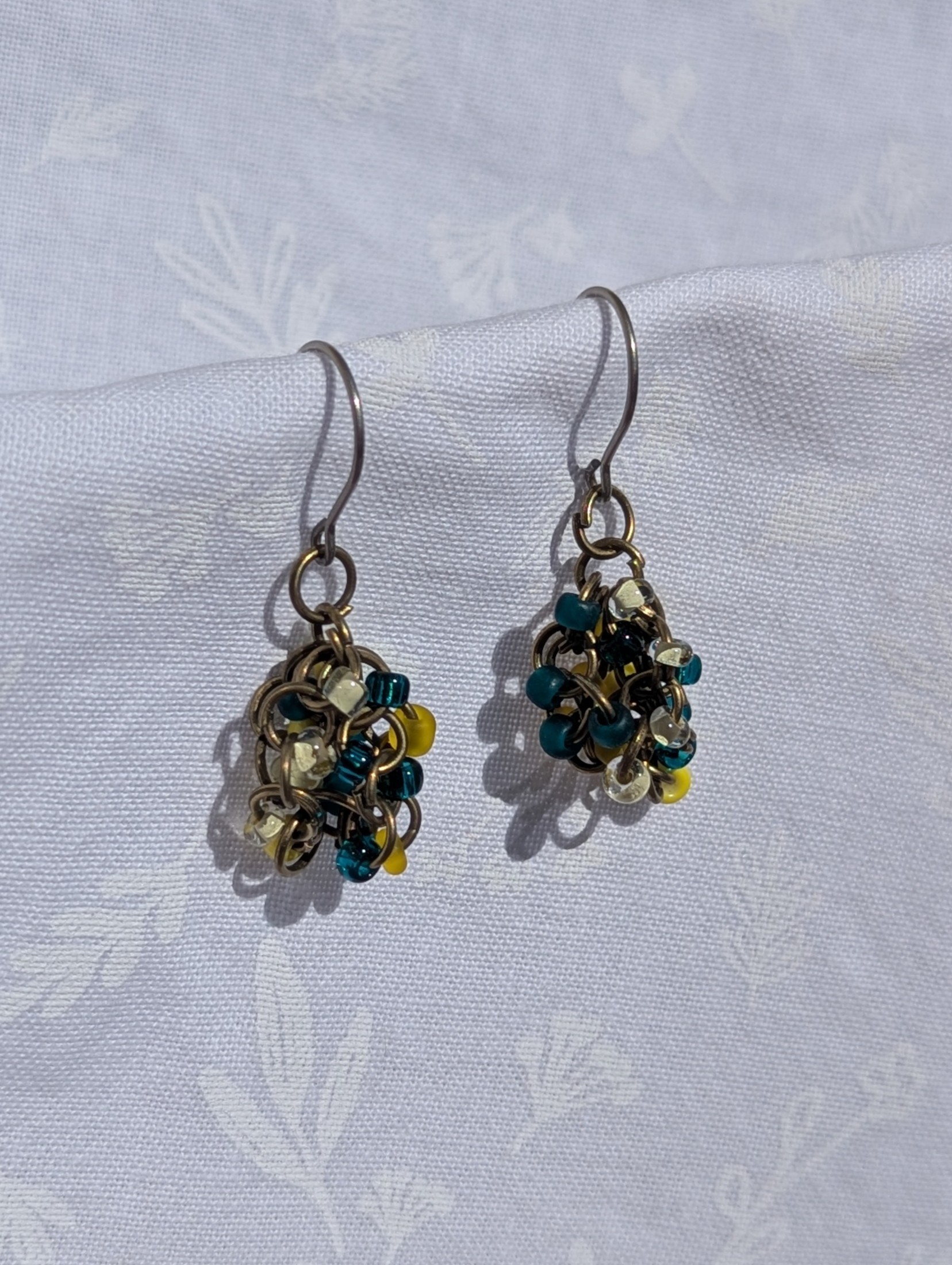 Ocean Sunrise Earrings: A pair of handmade beaded earrings featuring teal, yellow, and clear glass seed beads in varying textures, attached to titanium hooks with pewter-colored jump rings