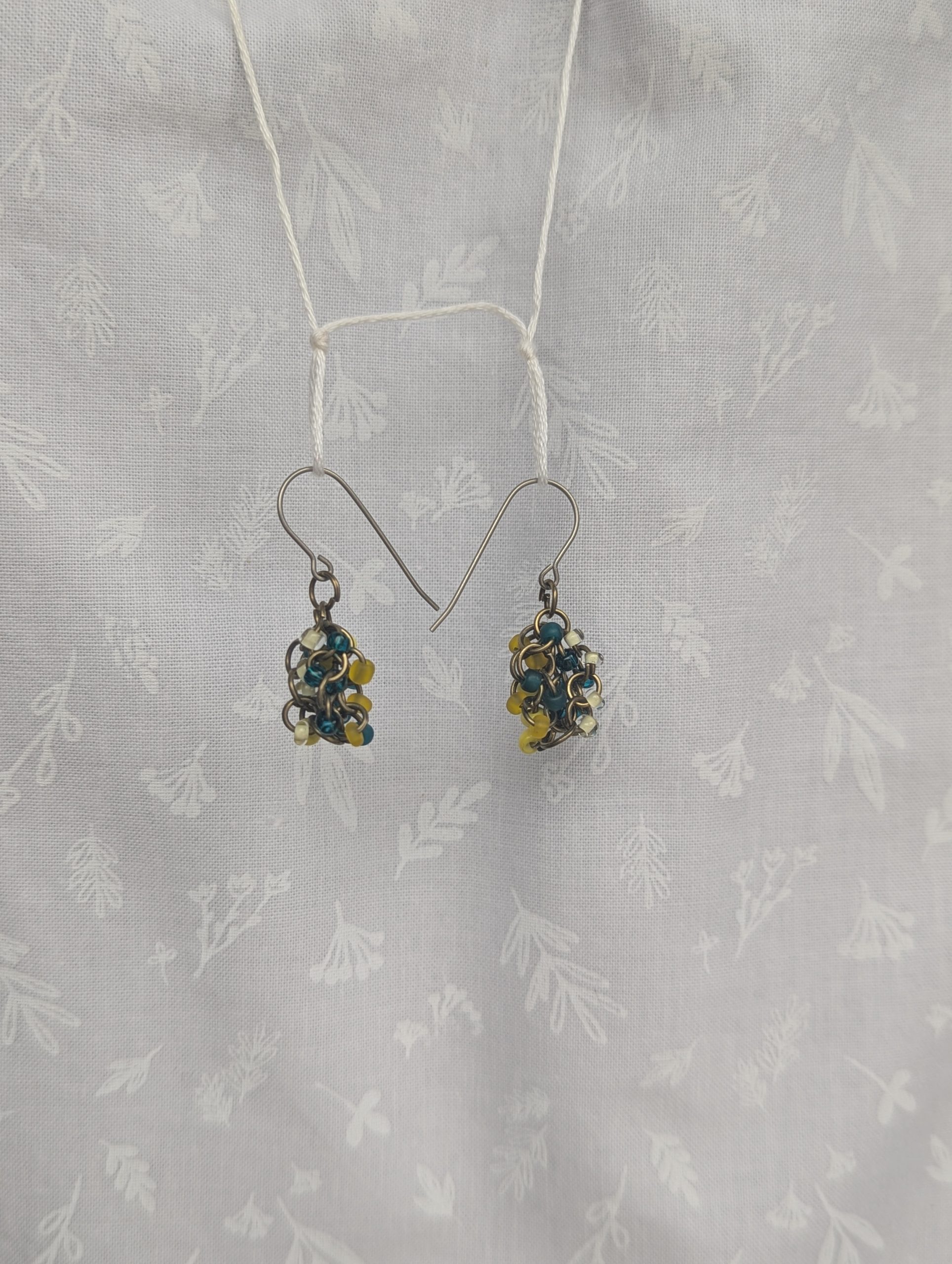 Ocean Sunrise Earring: A pair of handmade beaded earrings featuring teal, yellow, and clear glass seed beads in varying textures, attached to titanium hooks with pewter-colored jump rings.