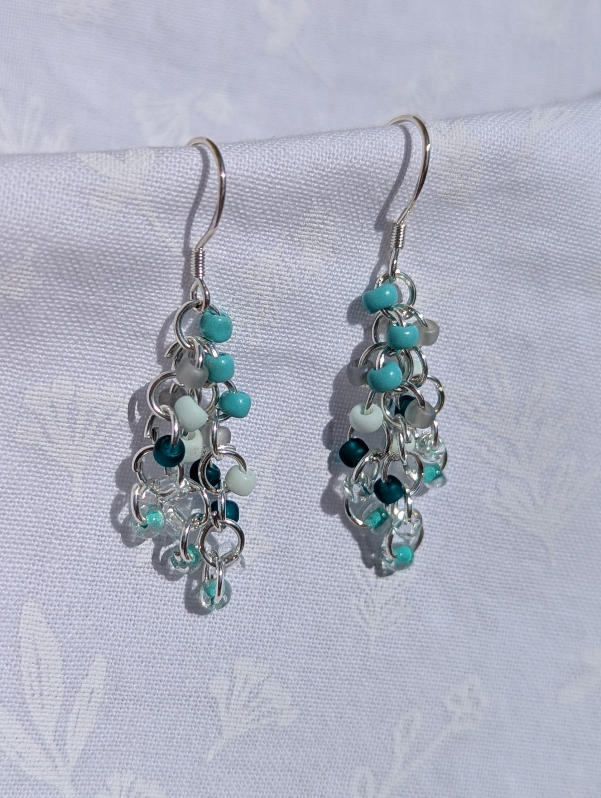 Coastal Breeze Earrings: Handmade beaded earrings featuring teal, seafoam green, and clear glass seed beads in varied textures, attached to sterling silver hooks with silver-colored jump rings