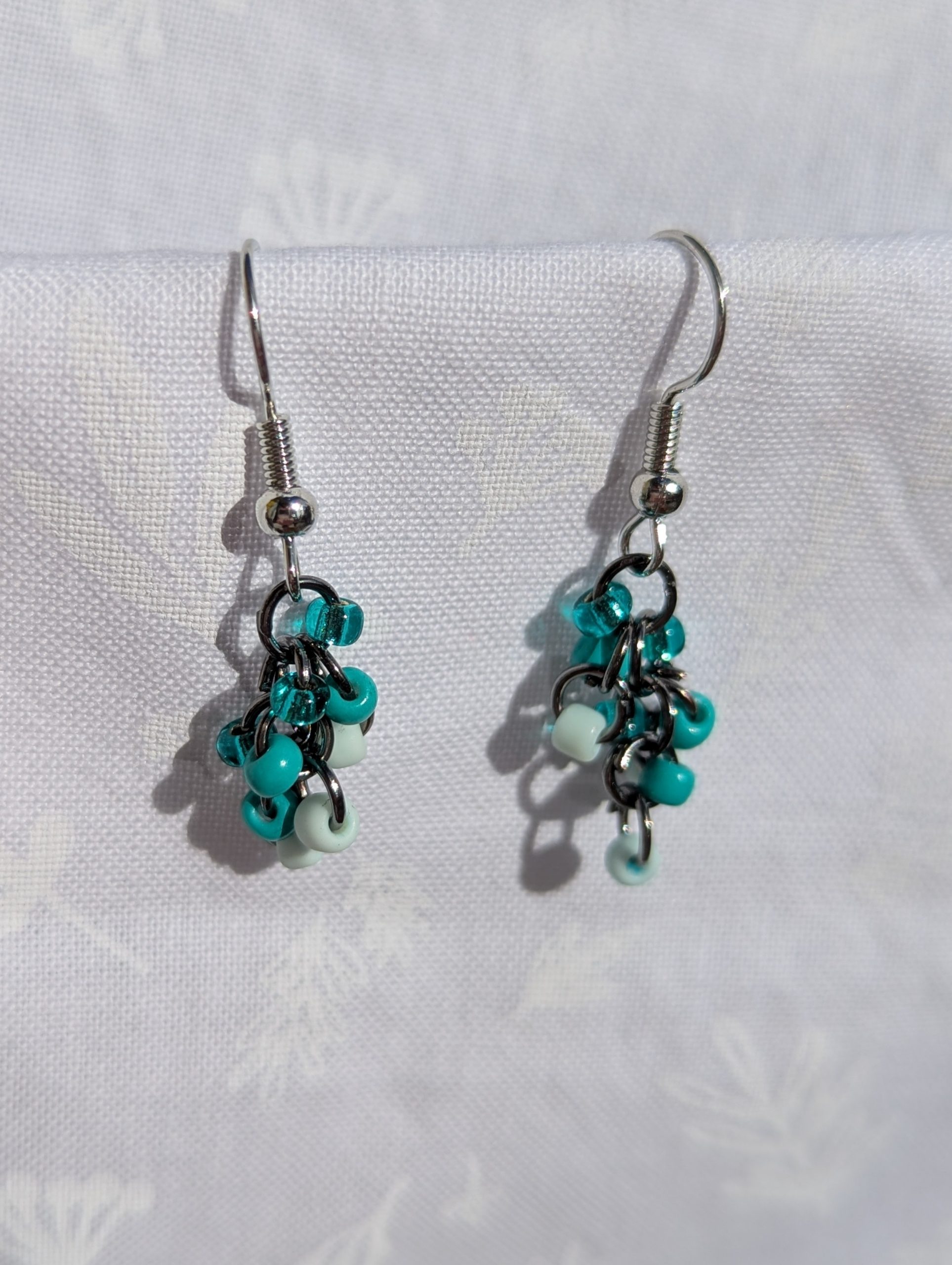 These elegant seafoam drops earrings feature nine seafoam and teal glass seed beads per earring in three shades. They are intricately linked with dark metal jump rings.