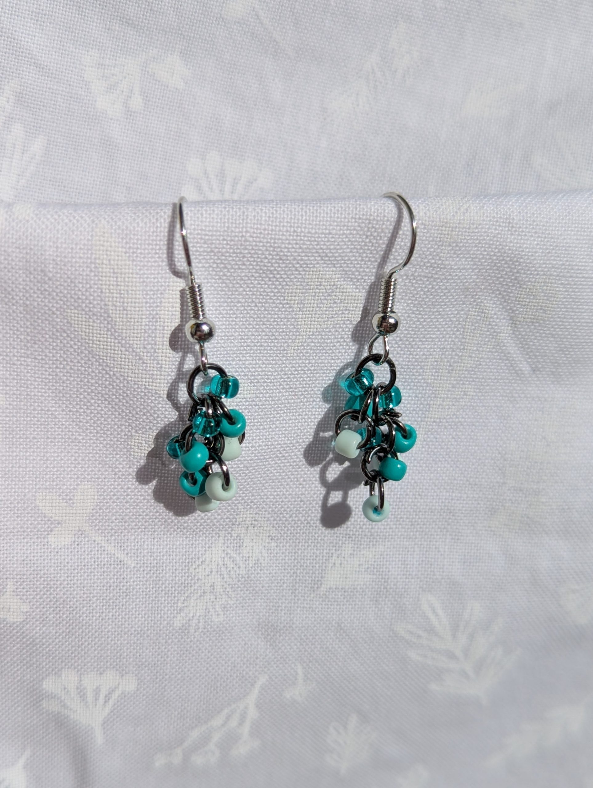 These elegant seafoam drops earrings feature nine seafoam and teal glass seed beads per earring in three shades. They are intricately linked with dark metal jump rings.