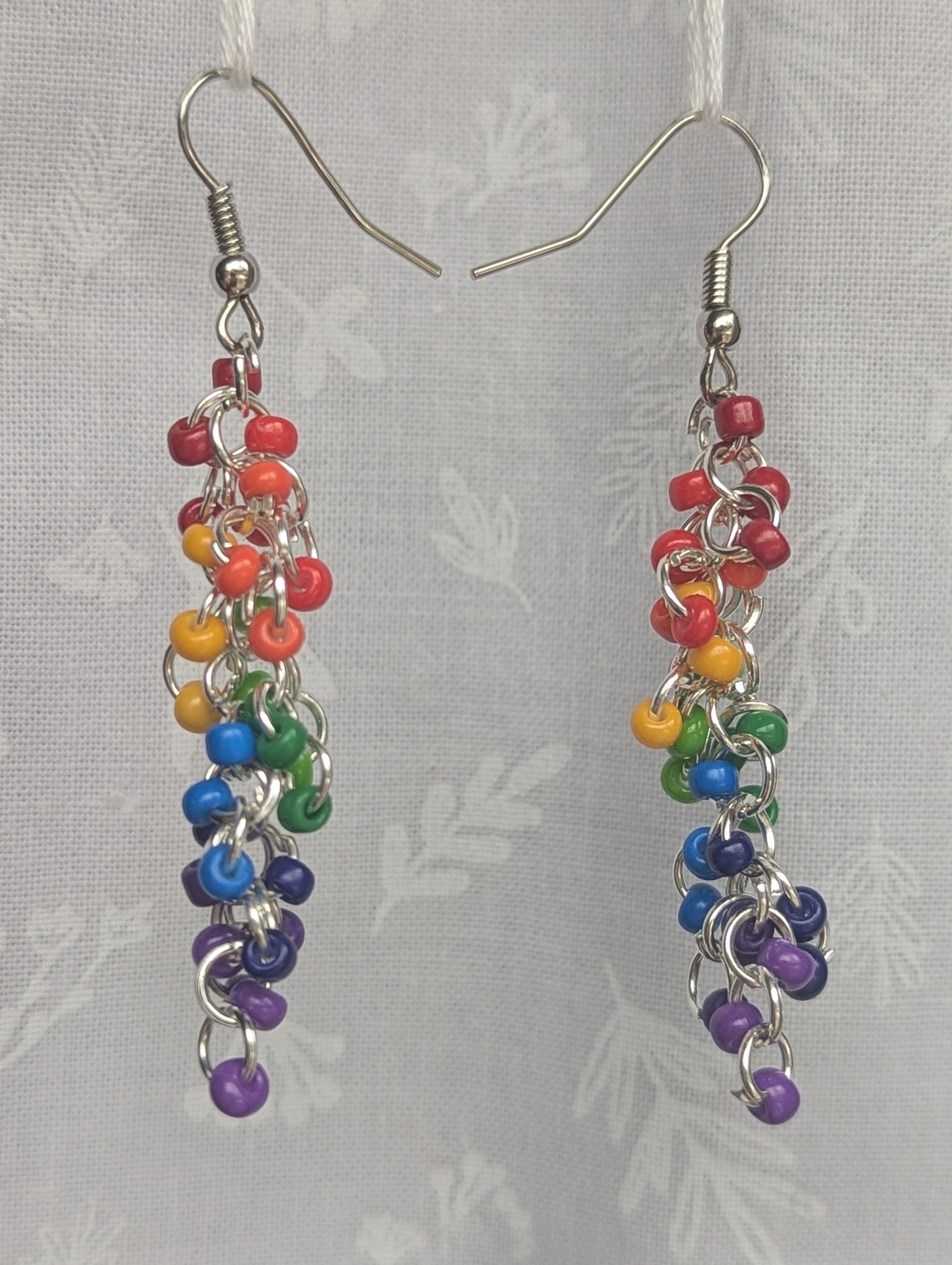 A pair of handmade rainbow dangle earrings featuring red, orange, yellow, green, blue, indigo, and purple glass seed beads with steel-colored hooks and jump rings.