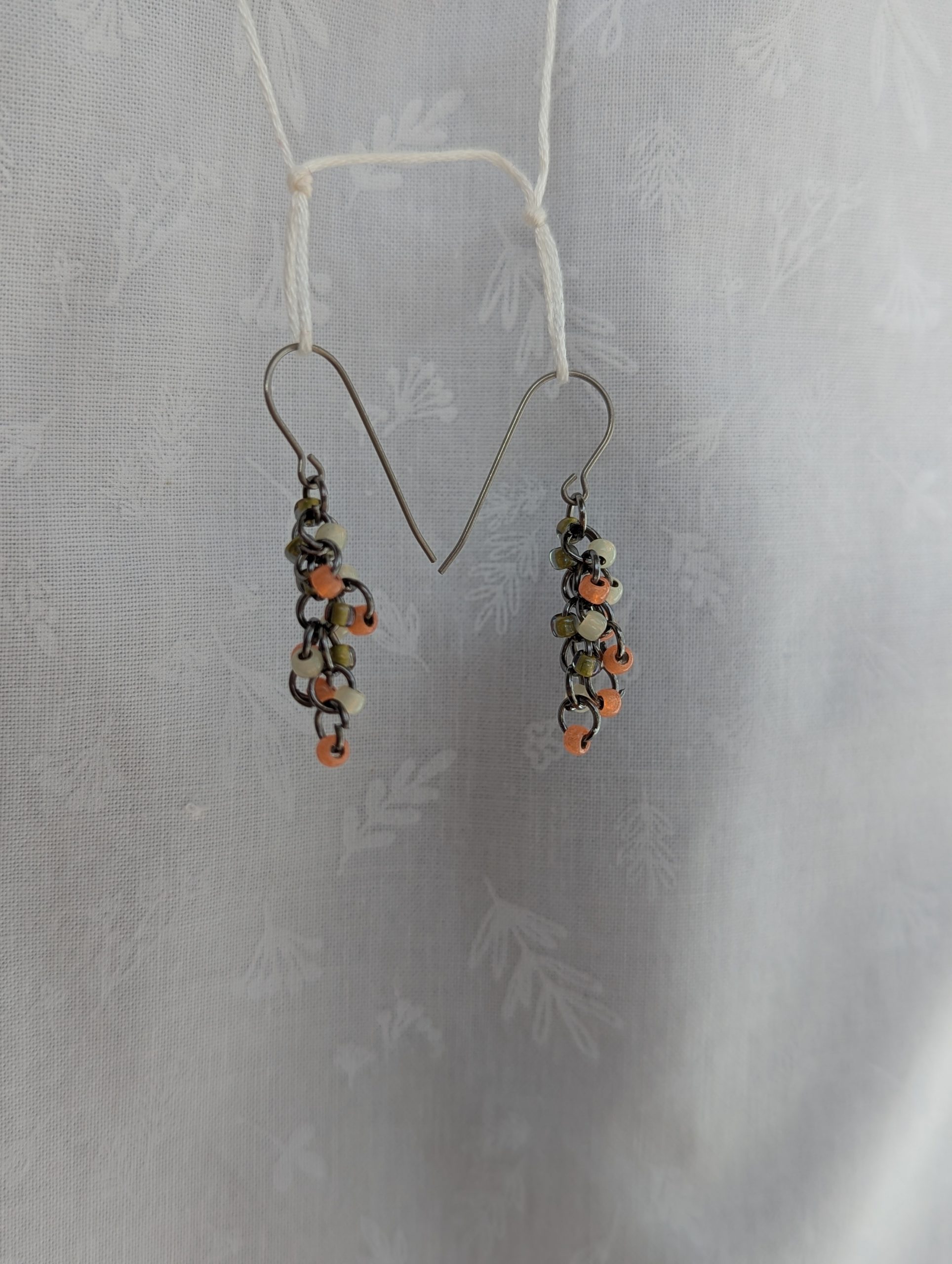 A pair of handcrafted dangle earrings featuring salmon, white, and green glass seed beads attached to pewter-colored jump rings, with hypoallergenic titanium hooks.