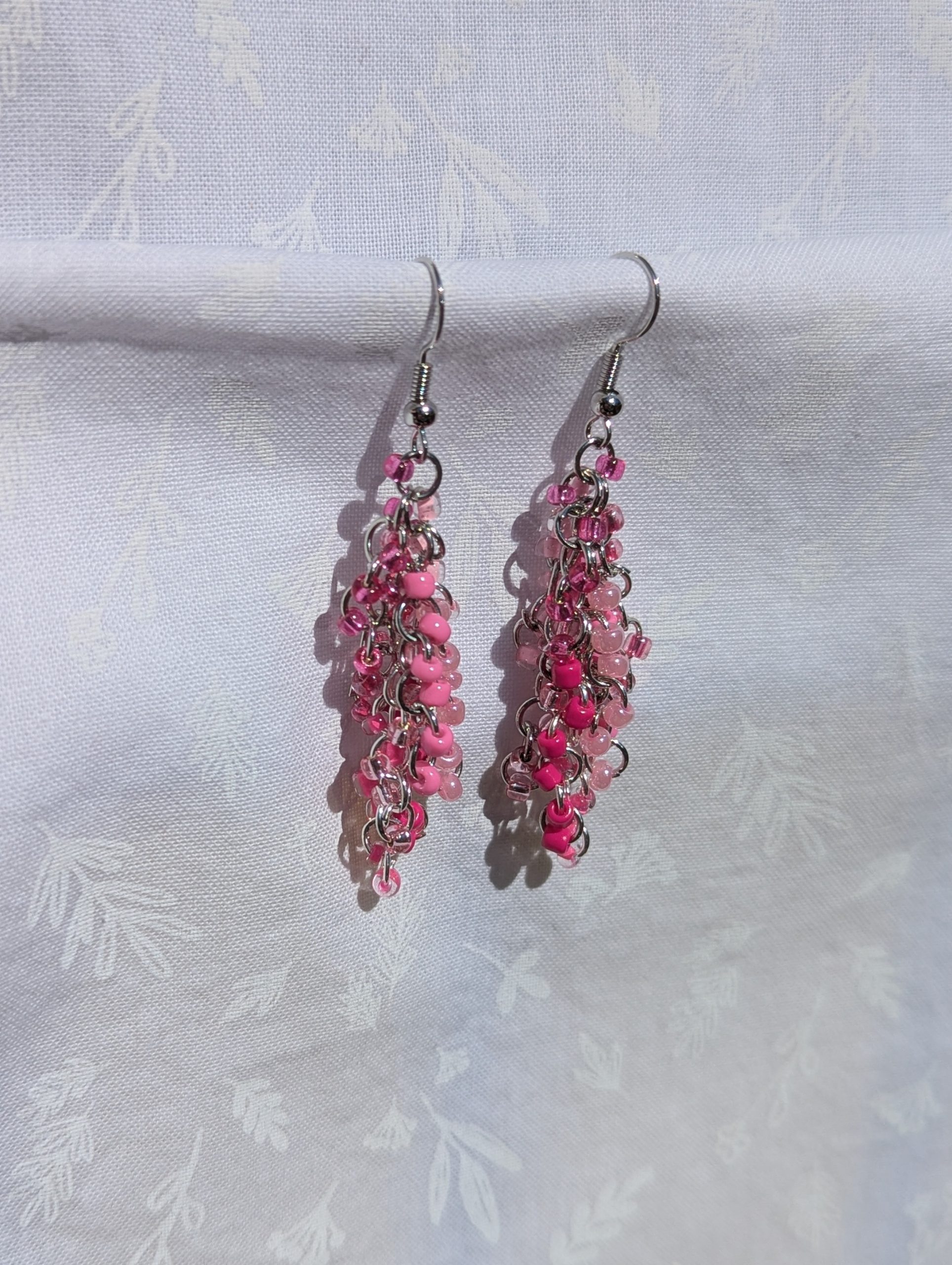 A pair of handmade pink beaded earrings with cascading glass seed beads in 8 shades of pink, linked with steel jump rings and hooks, displayed against a white fabric background
