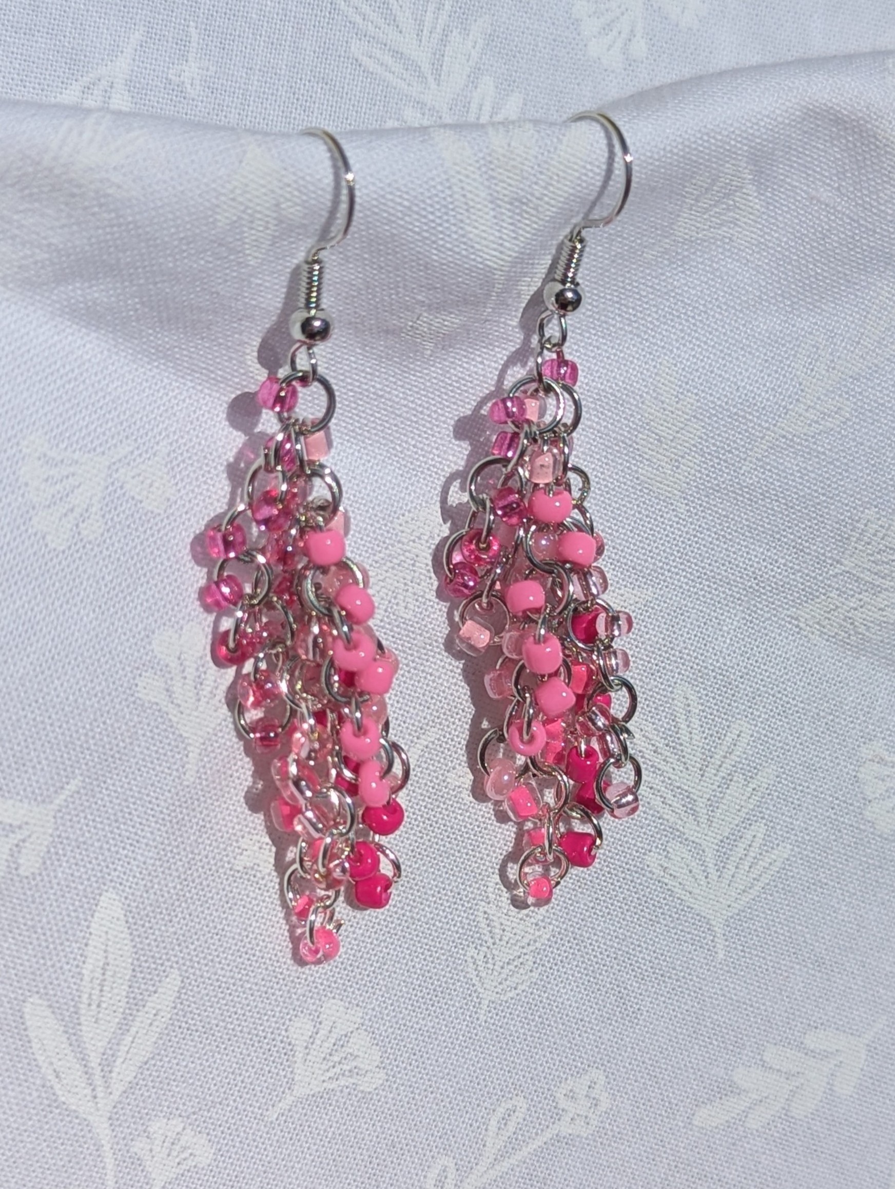 Pink Waterfall Earrings: A pair of handmade pink beaded earrings with cascading glass seed beads in 8 shades of pink, linked with steel jump rings and hooks, displayed against a white fabric background
