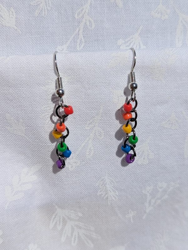 A pair of handmade mini rainbow earrings featuring six colorful glass seed beads—red, orange, yellow, green, blue, and purple—linked with pewter-colored jump rings and silver-colored earring hooks, displayed against a white fabric background.