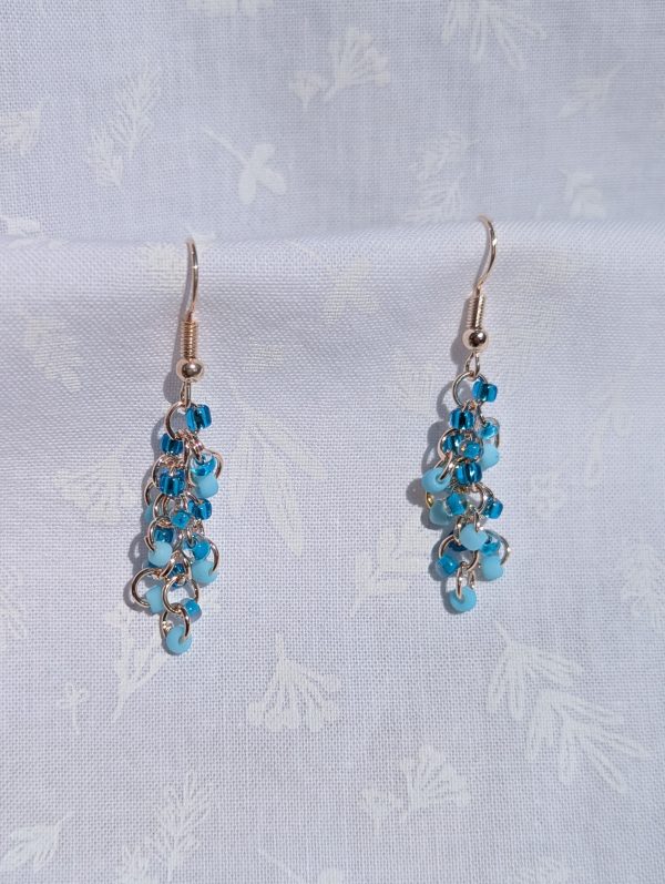Clear Blue Sky Earrings: A pair of handmade earrings featuring three shades of blue glass seed beads, connected by rose-gold-colored jump rings and hooks in a cascading design.