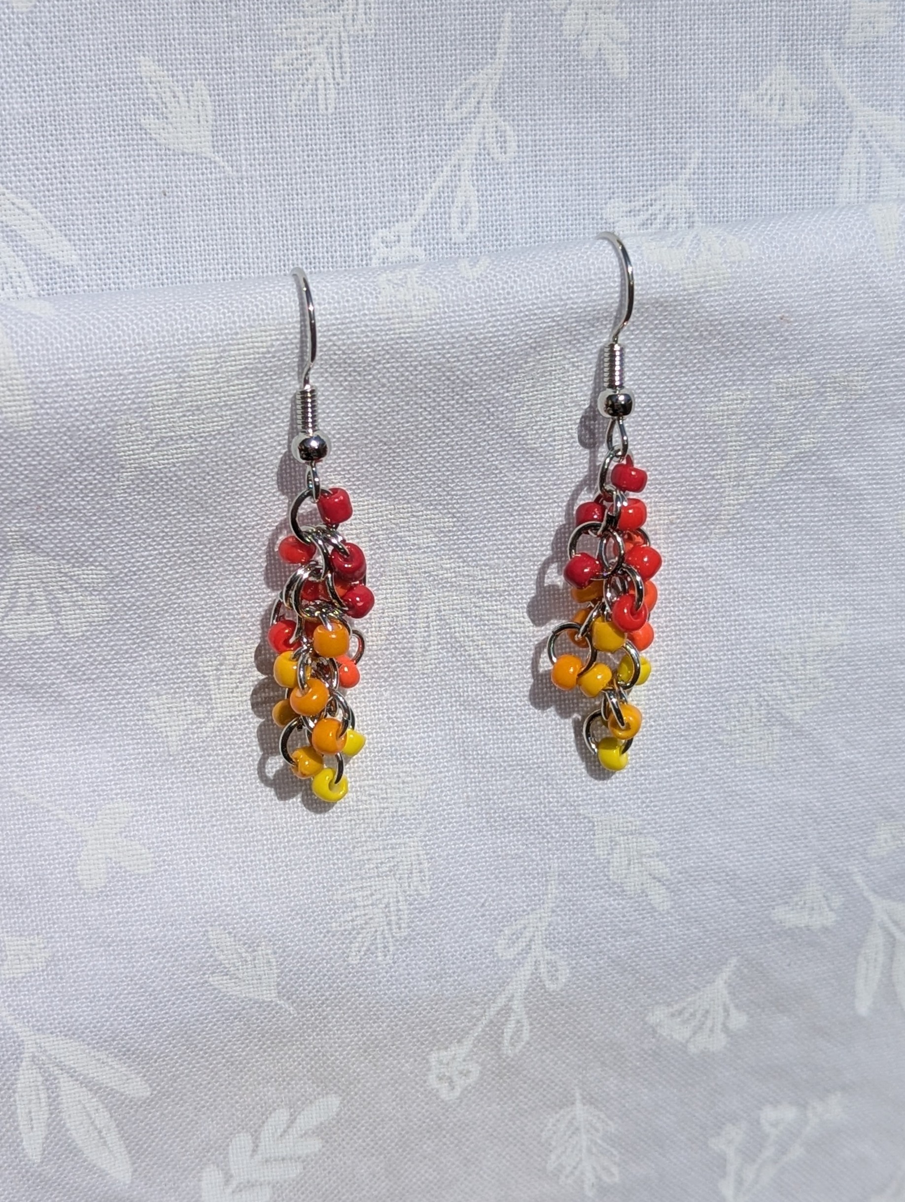 A pair of handmade earrings featuring cascading fuchsia, red, orange, and yellow glass seed beads, connected by steel-colored jump rings and hooks.