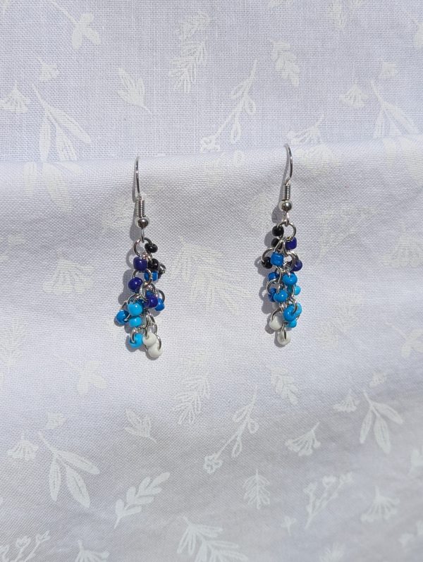 A pair of handmade earrings featuring cascading black, blue, and white glass seed beads, connected by steel-colored jump rings and hooks.