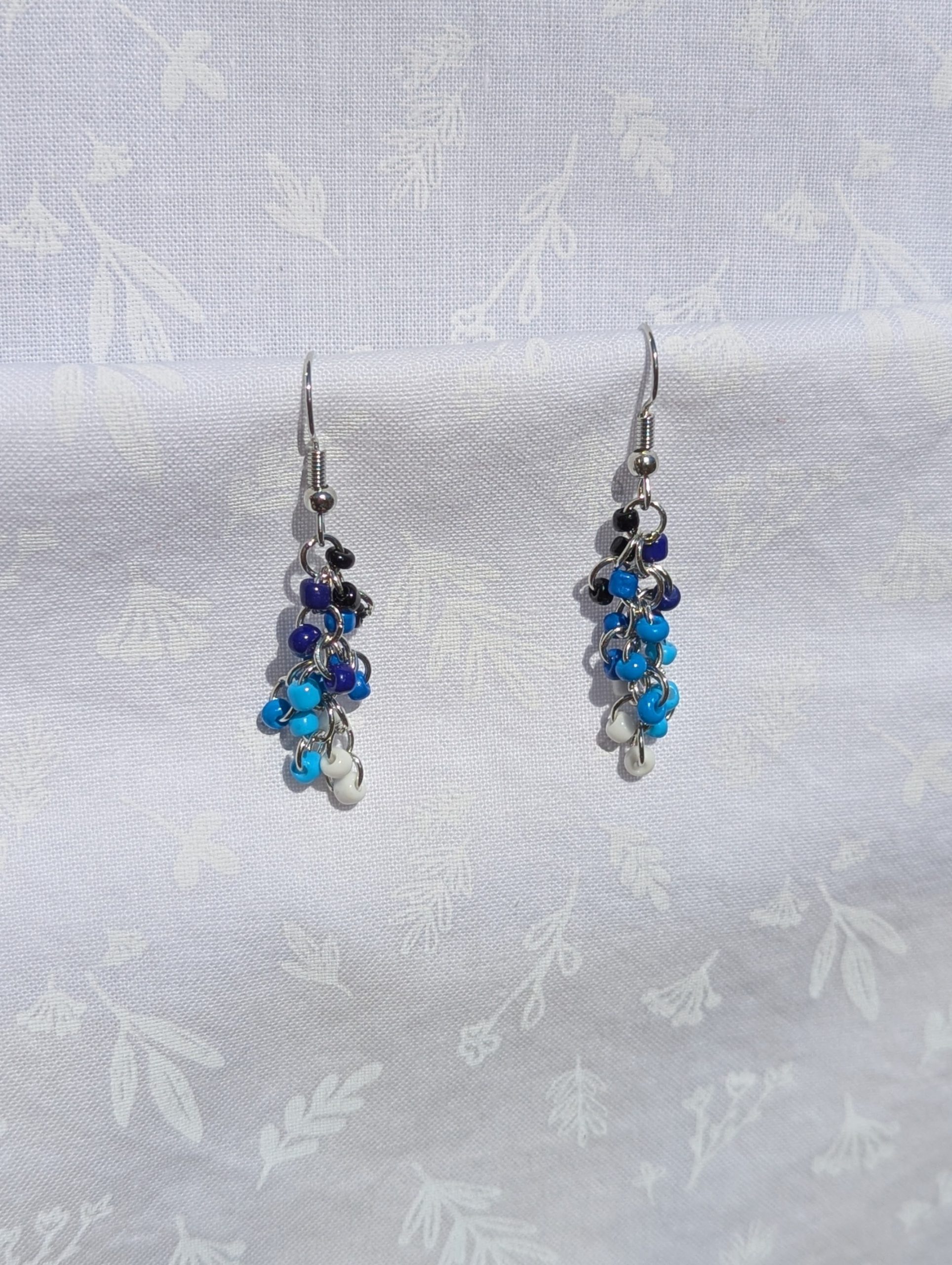 A pair of handmade earrings featuring cascading black, blue, and white glass seed beads, connected by steel-colored jump rings and hooks.