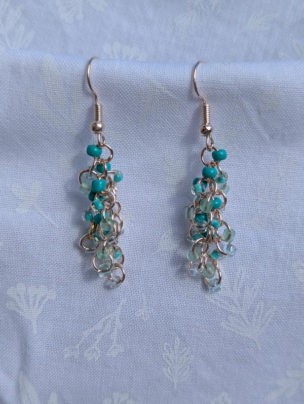 The Seafoam green and teal earrings feature a blend of teal glass seed beads of various hues each with a variety of texture. A total of 18 beads per earring—three beads per hue—cascade gracefully, creating a vibrant and eye-catching effect.