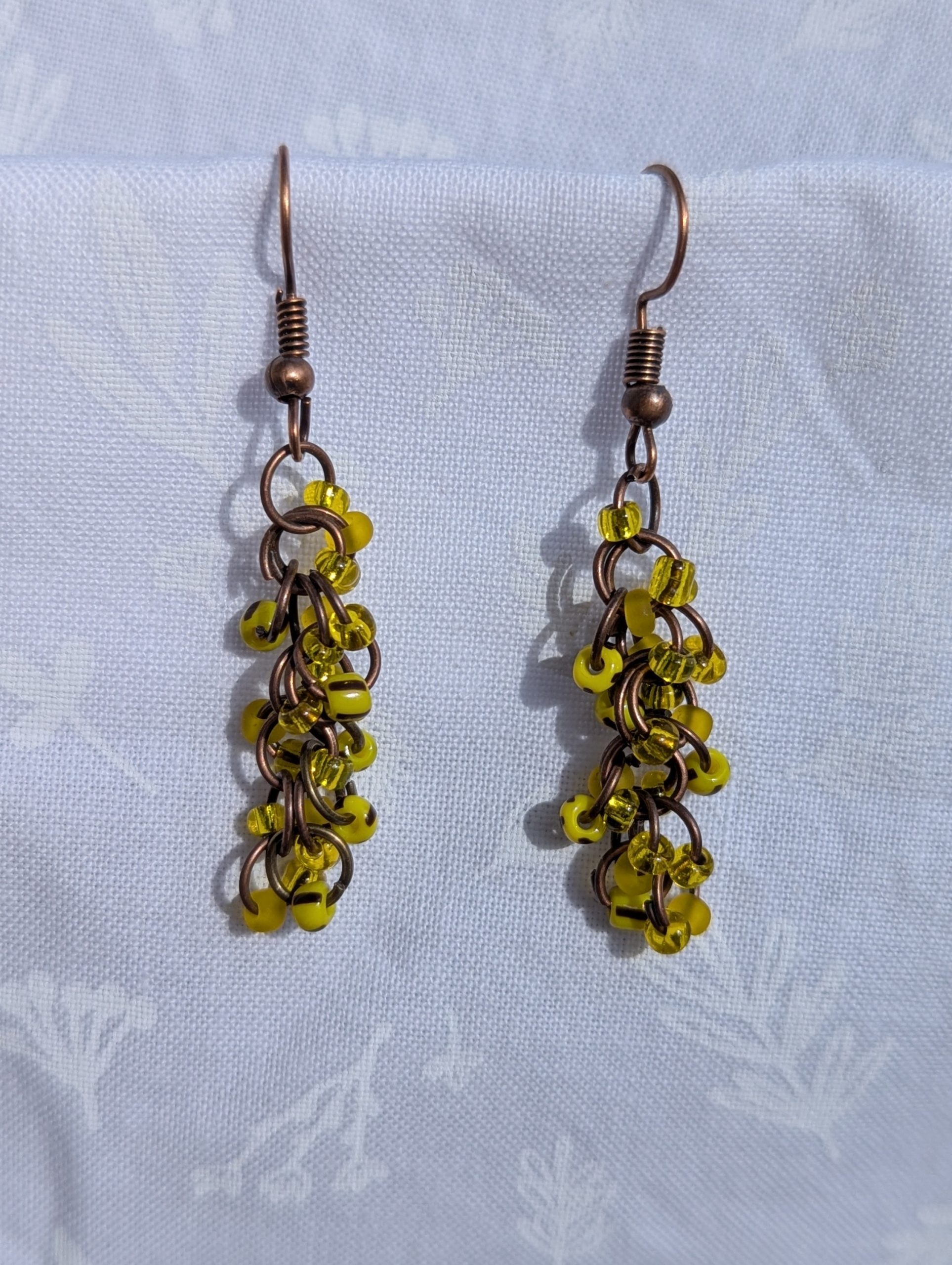 Golden Glow Cascade Earrings: A pair of handmade yellow beaded earrings with antique bronze hooks and jump rings, displayed against a soft, floral-patterned fabric background.