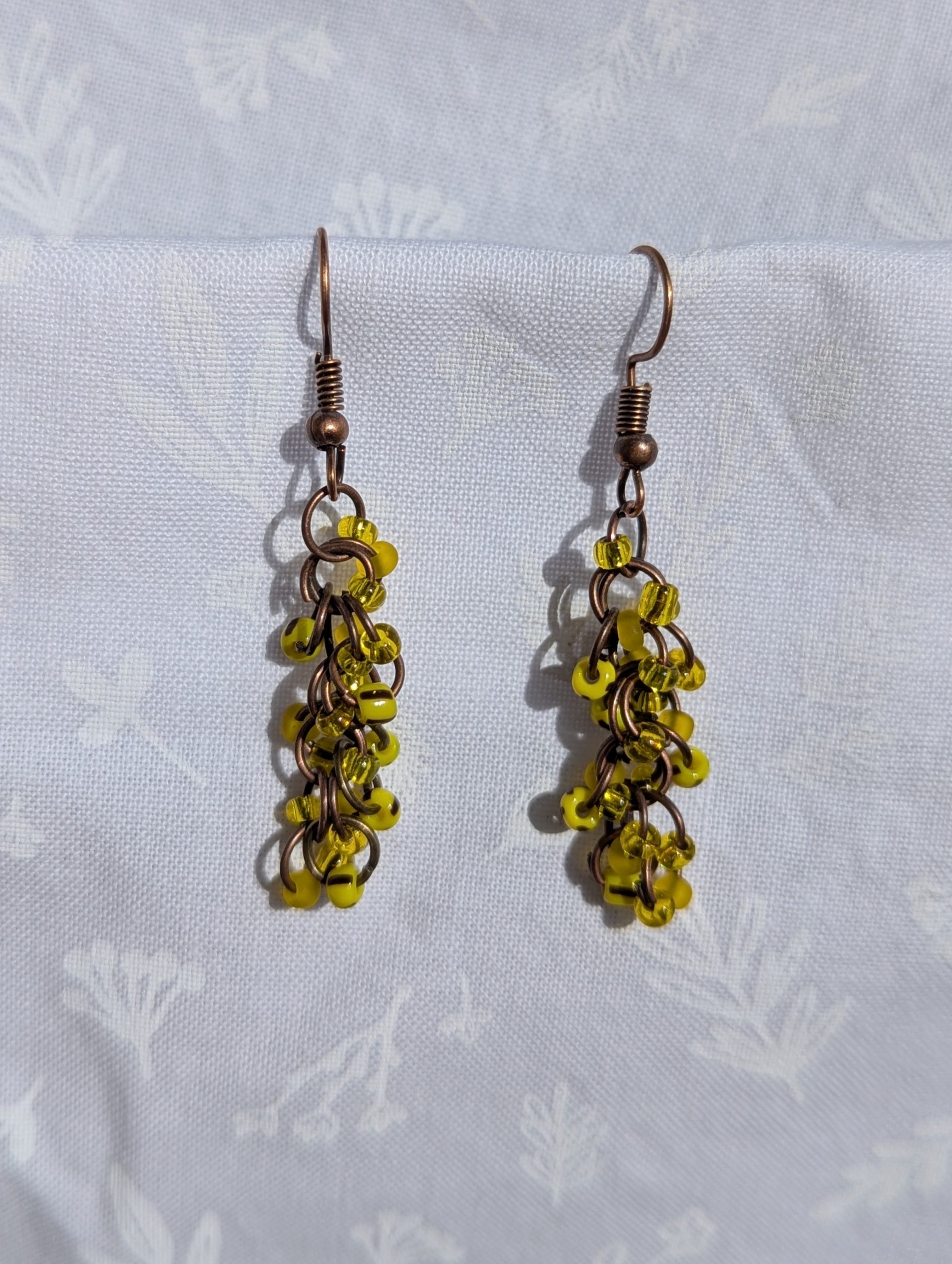 Golden Glow Cascade Earrings: A pair of handmade yellow beaded earrings with antique bronze hooks and jump rings, displayed against a soft, floral-patterned fabric background.