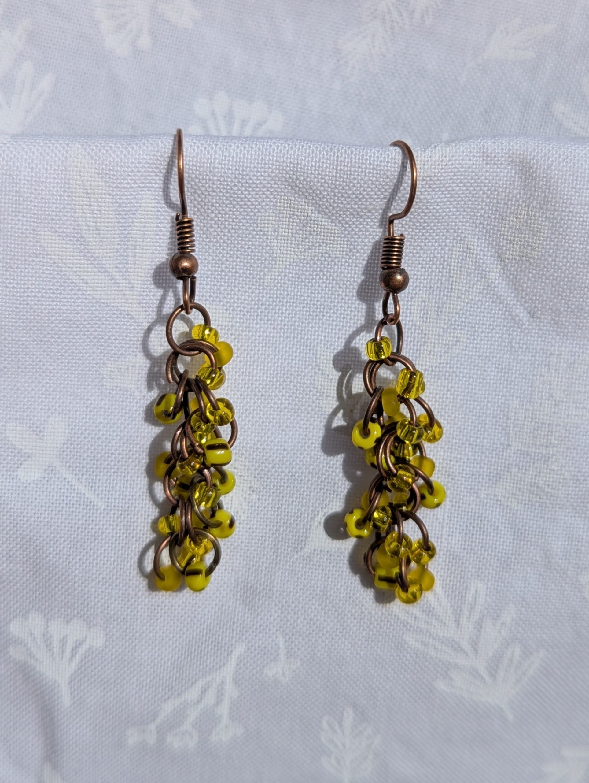 Golden Glow Cascade Earrings: A pair of handmade yellow beaded earrings with antique bronze hooks and jump rings, displayed against a soft, floral-patterned fabric background.