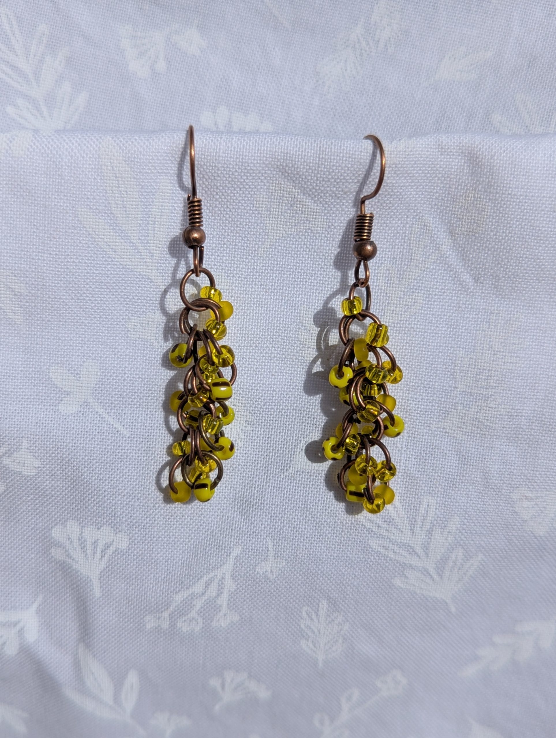 Golden Glow Cascade Earrings: A pair of handmade yellow beaded earrings with antique bronze hooks and jump rings, displayed against a soft, floral-patterned fabric background.