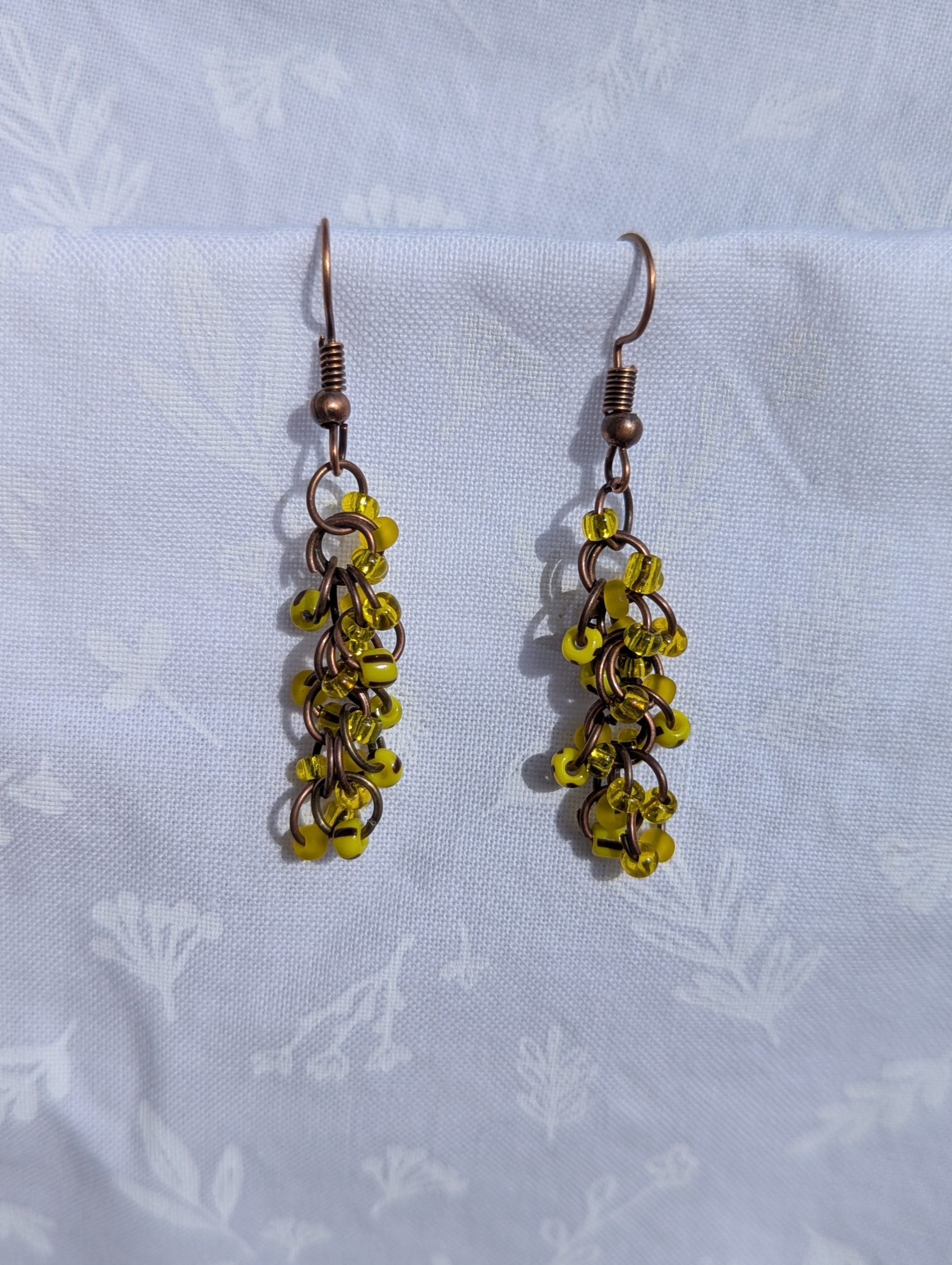 Golden Glow Cascade Earrings: A pair of handmade yellow beaded earrings with antique bronze hooks and jump rings, displayed against a soft, floral-patterned fabric background.