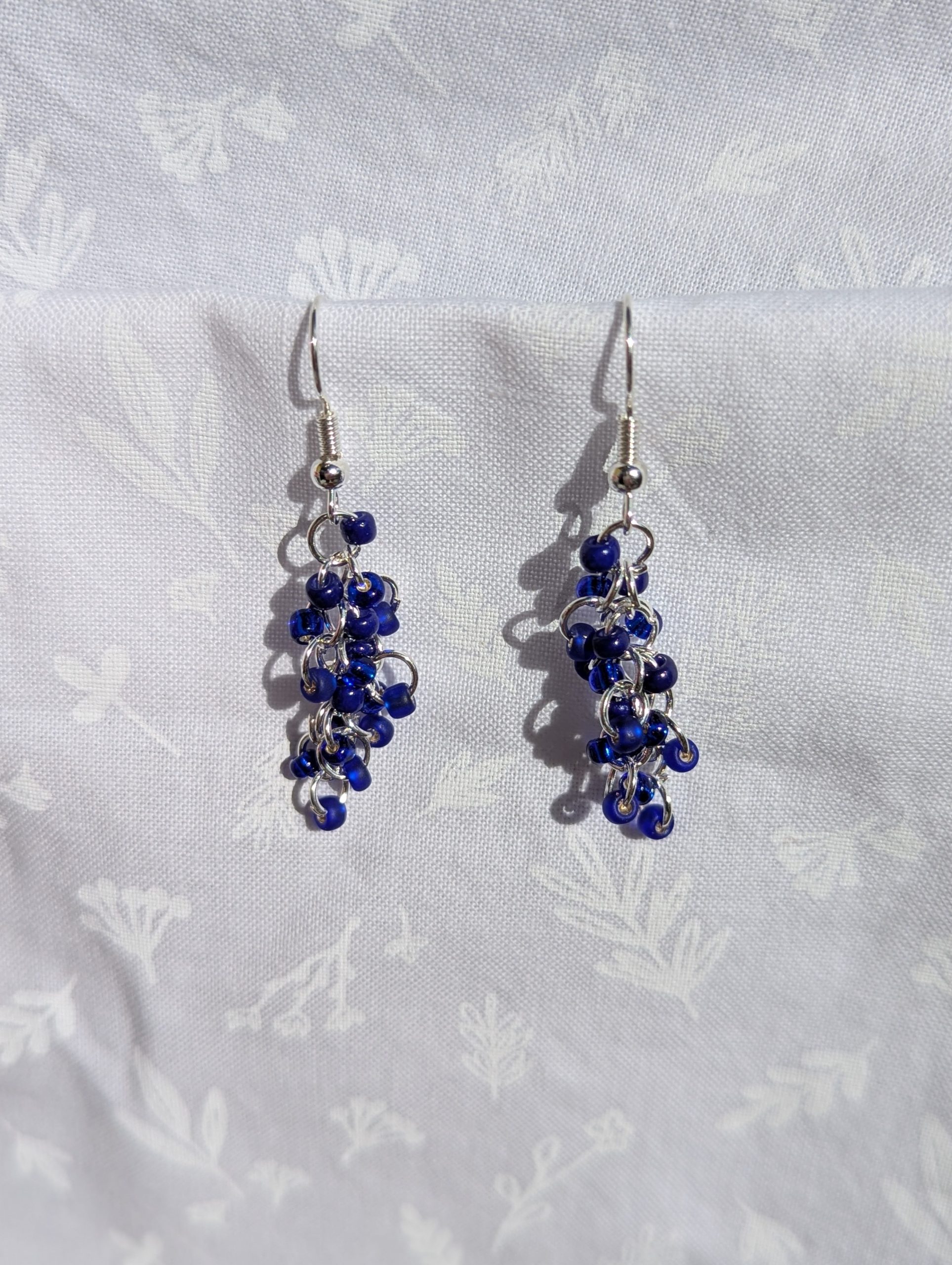 Midnight Cascade Earrings: A pair of handmade earrings featuring cascading deep blue glass seed beads connected by silver-toned jump rings and hooks, displayed against a soft white fabric background.