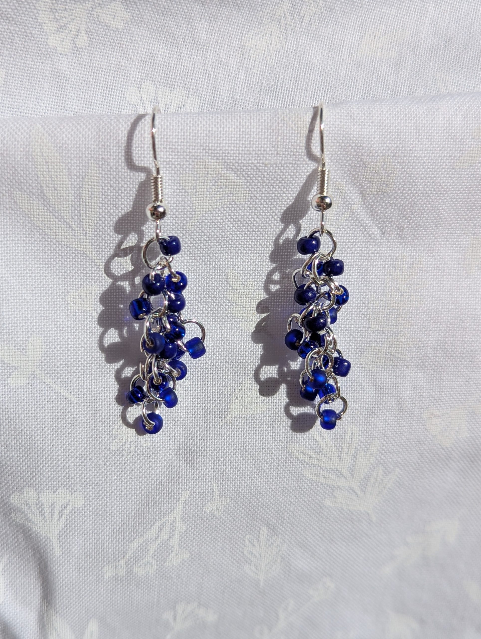 Midnight-Cascade earrings: A pair of handmade earrings featuring cascading deep blue glass seed beads connected by silver-toned jump rings and hooks, displayed against a soft white fabric background.