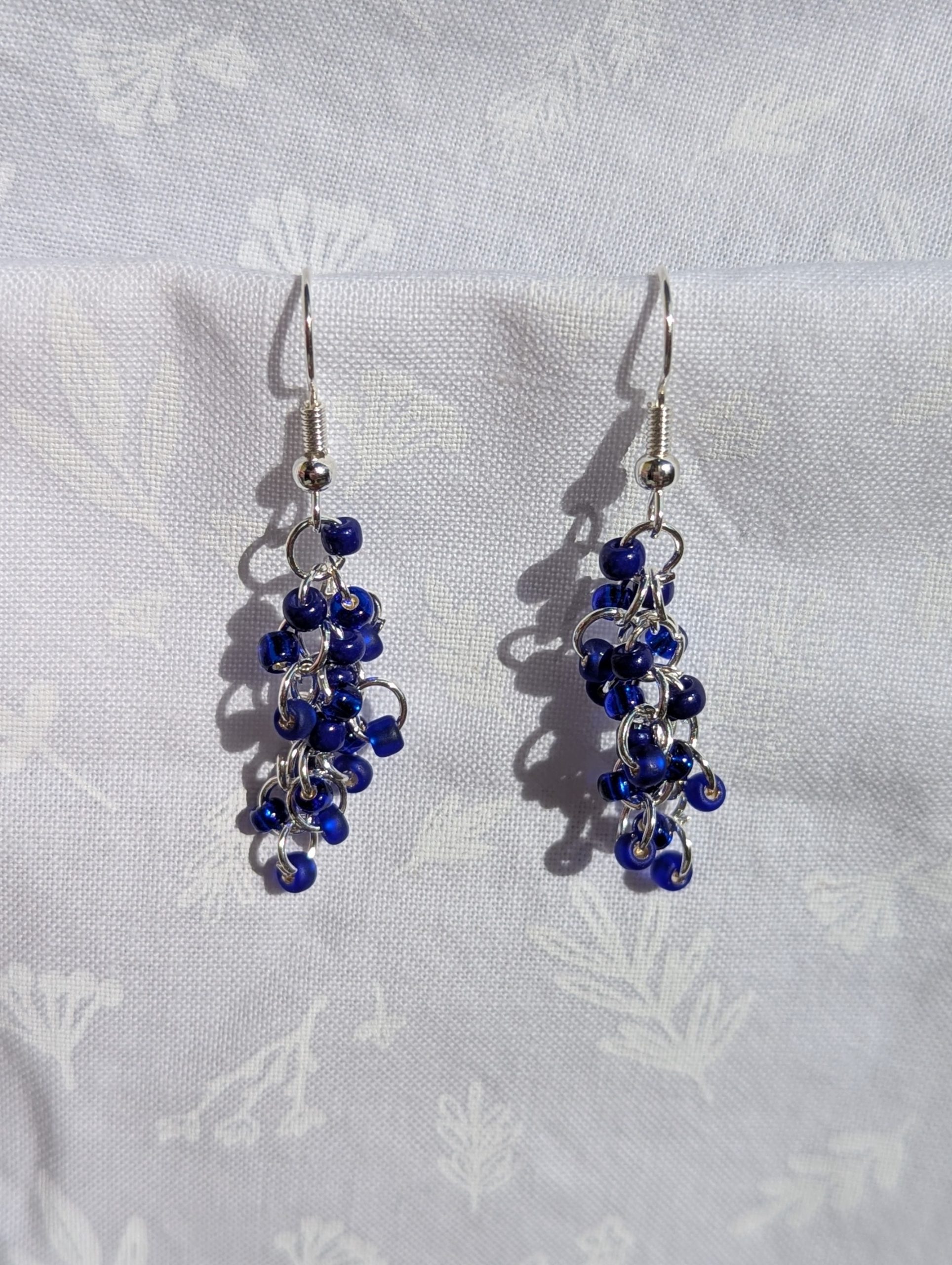 Midnight Cascade Earrings: A pair of handmade earrings featuring cascading deep blue glass seed beads connected by silver-toned jump rings and hooks, displayed against a soft white fabric background.