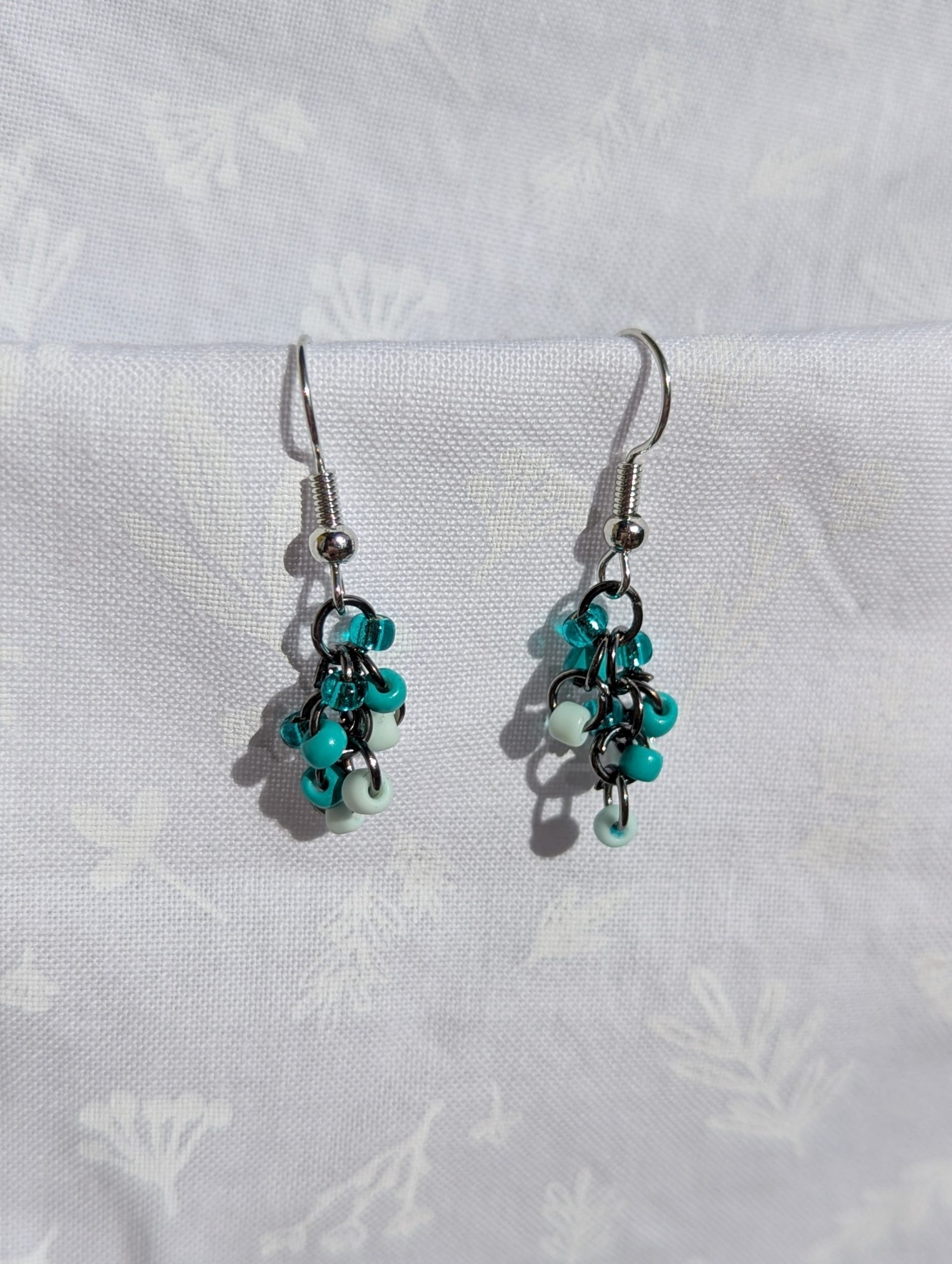 These elegant seafoam drops earrings feature nine seafoam and teal glass seed beads per earring in three shades. They are intricately linked with dark metal jump rings.