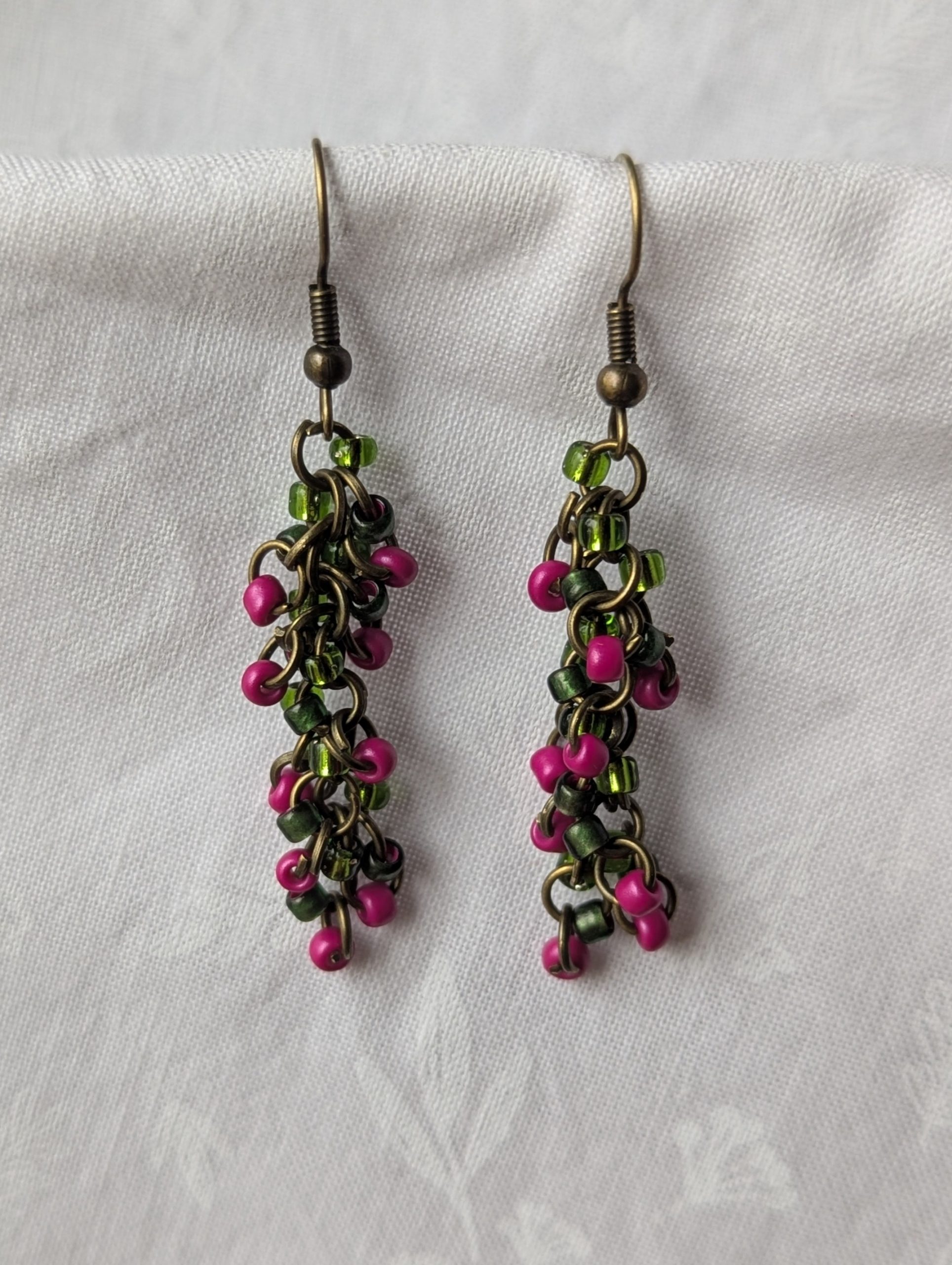 Enchanted Garden Earrings: Handmade earrings featuring deep green, deep fuchsia, and fuchsia-lined green beads, intricately connected with bronze jump rings and matching hooks.