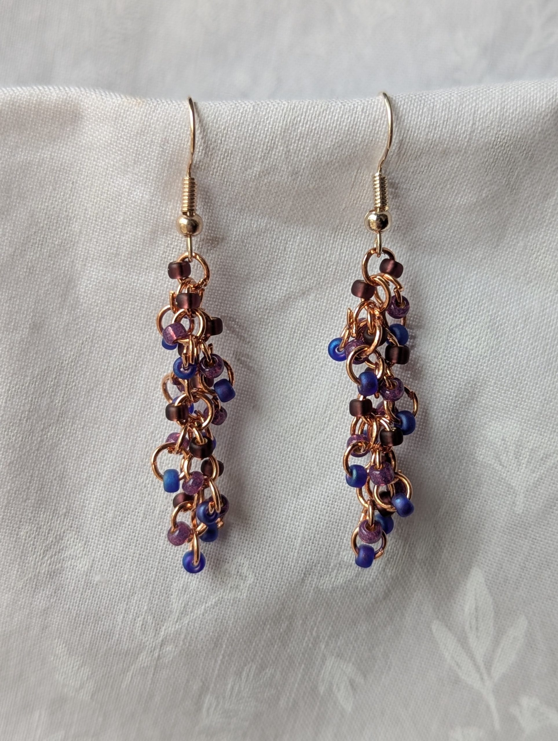 Dragon Scales Earrings featuring iridescent blue/purple, matte deep purple, and pearly amethyst beads, connected with rose-gold jump rings and matching hooks.