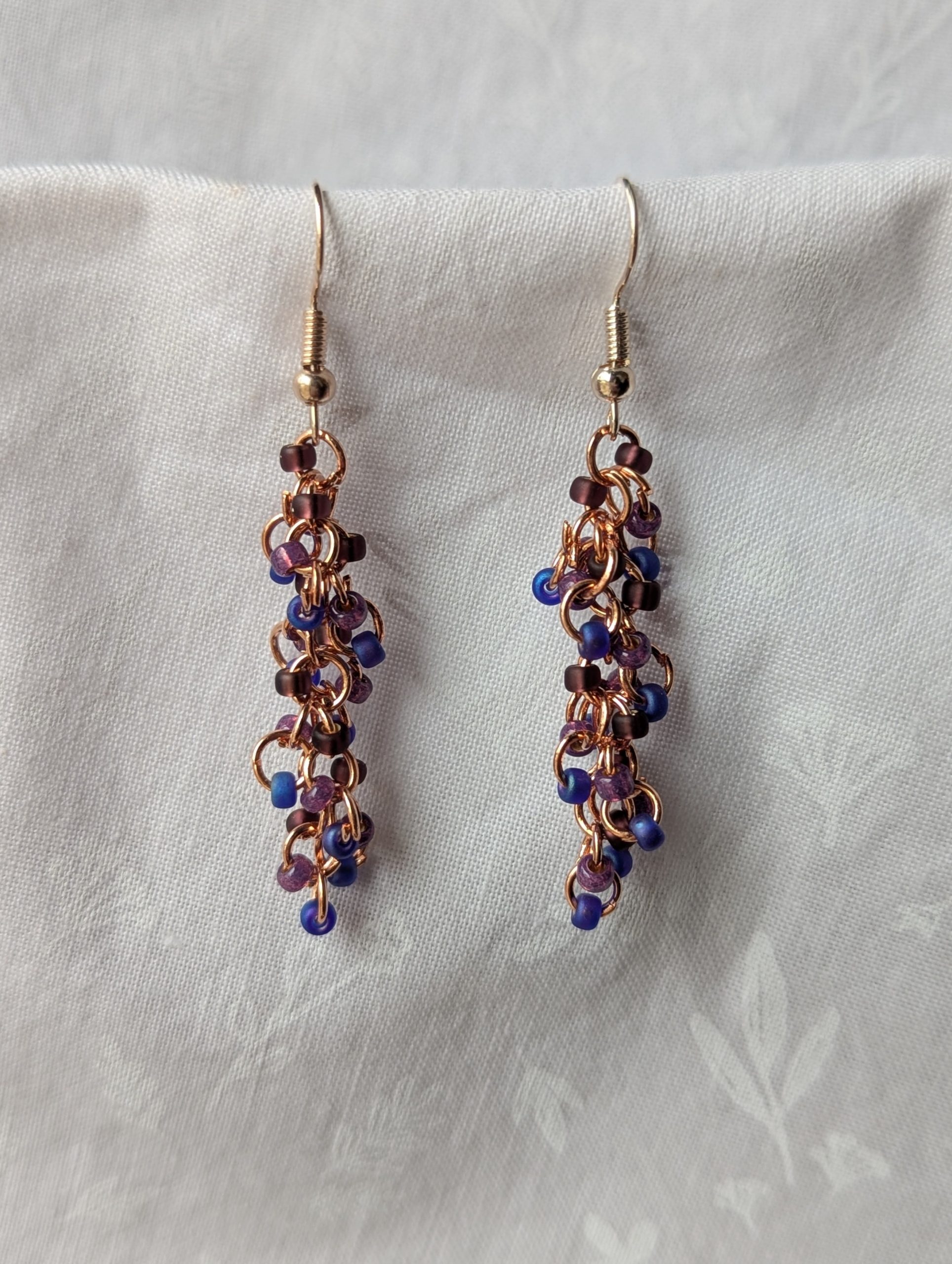 Dragon Scales Earrings featuring iridescent blue/purple, matte deep purple, and pearly amethyst beads, connected with rose-gold jump rings and matching hooks.
