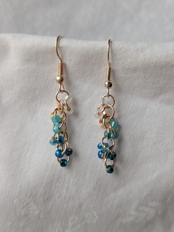 Mermaid Tears earrings with 12 beads in ocean tones: pearly white, shiny cream, pearly cyan, translucent ocean blue, metallic blue, and shiny teal, connected with rose-gold jump rings and hooks.