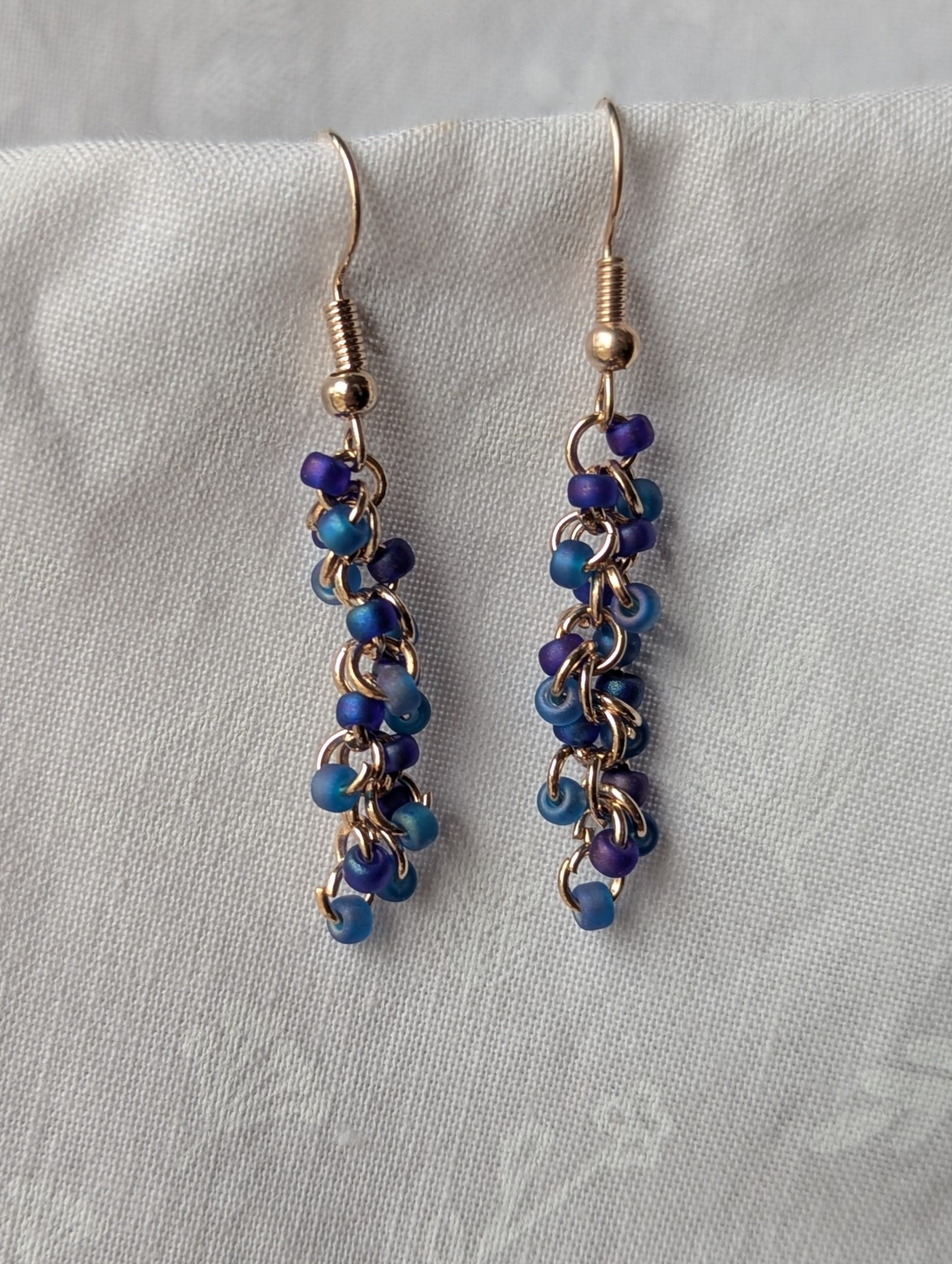 Stormy Waves earrings with 18 beads—nine iridescent blue and nine iridescent purple—linked with rose-gold jump rings and matching earring hooks.
