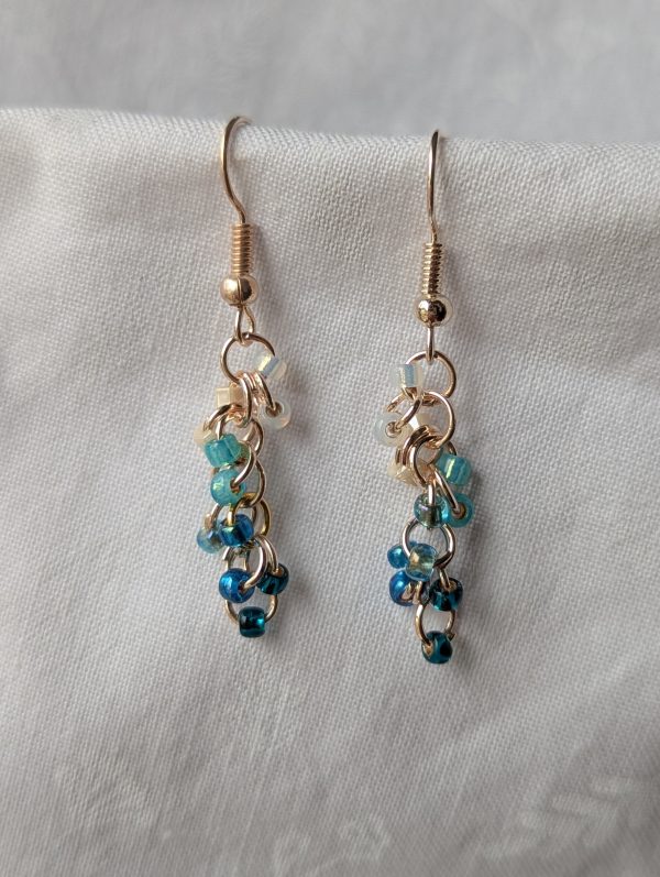 Ocean Tide earrings with 14 beads in seven ocean-inspired colors: clear, matte gray, pearly blue/gray, iridescent blue, metallic blue, matte deep blue, and solid indigo, connected with gold-colored jump rings and hooks.