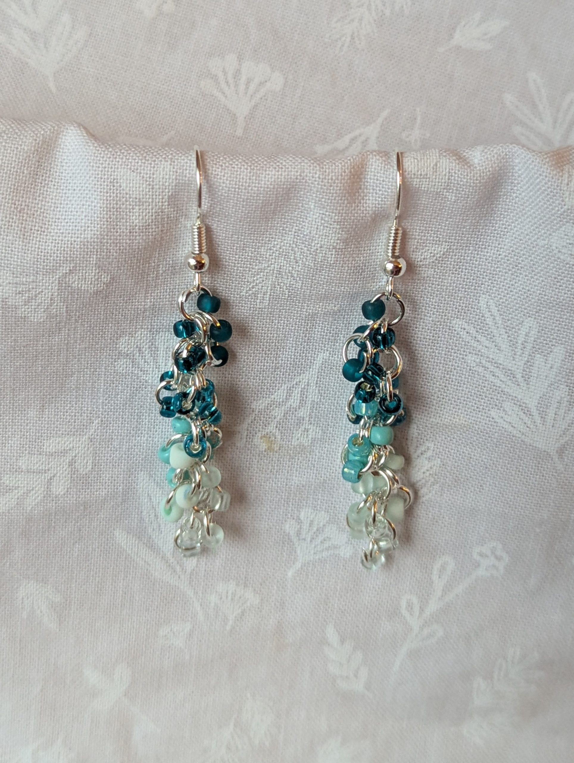 Caribbean Sea earrings with 27 beads in nine teal and cyan shades, including frosted, pearly, and translucent finishes. Connected with silver-colored jump rings and earring hooks.