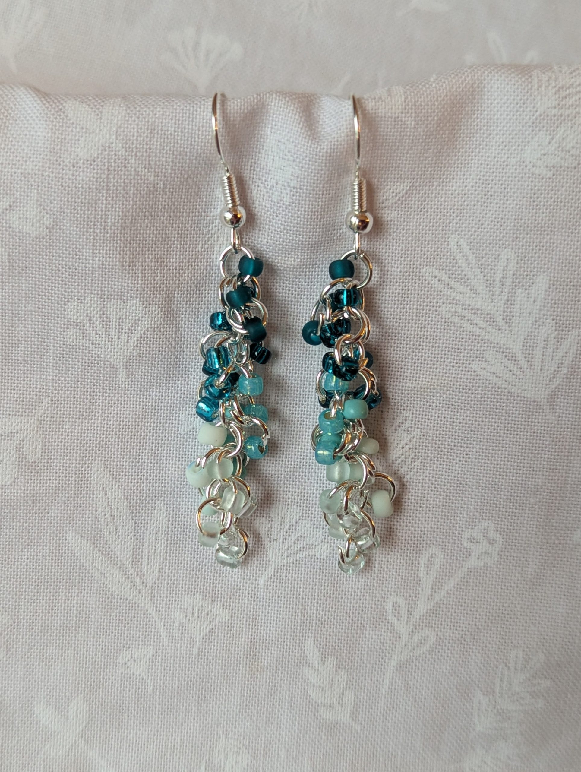 Caribbean Sea earrings with 27 beads in nine teal and cyan shades, including frosted, pearly, and translucent finishes. Connected with silver-colored jump rings and earring hooks.