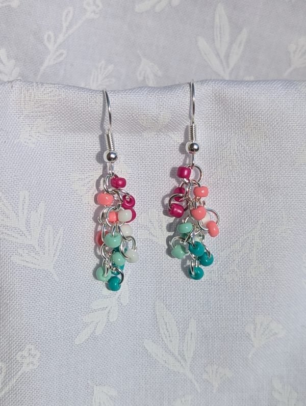 Tropical Breeze Earrings: Handmade earrings featuring 15 glass seed beads in teal, seafoam green, white, salmon pink, and fuchsia, arranged on silver-colored jump rings with matching earring hooks.