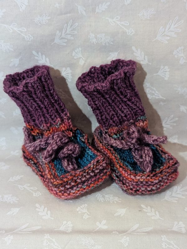 Multicolored baby booties in pink, purple, and turquoise with ribbed cuffs and soft pink ties, handknit for newborns.