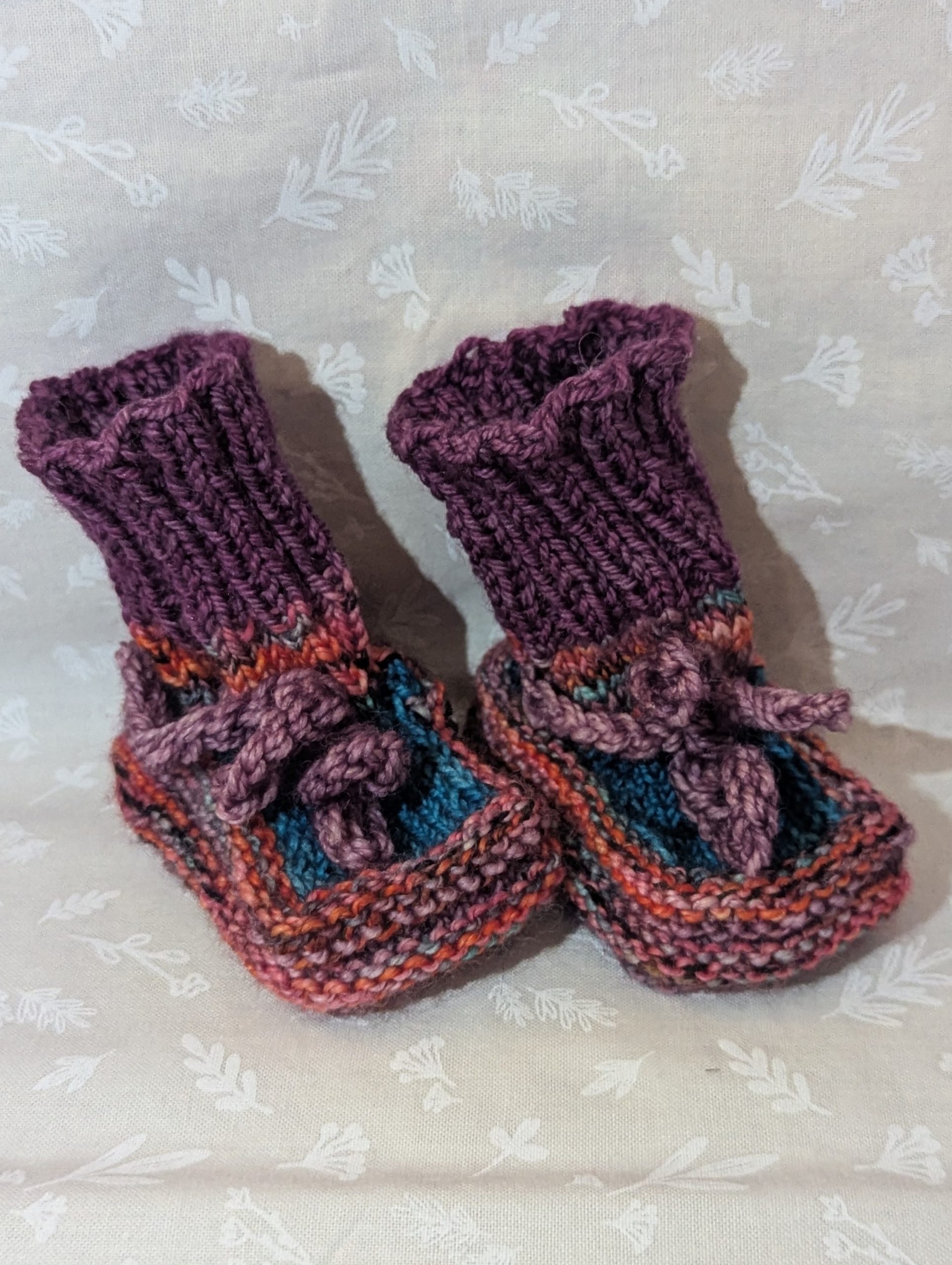 Multicolored baby booties in pink, purple, and turquoise with ribbed cuffs and soft pink ties, handknit for newborns.
