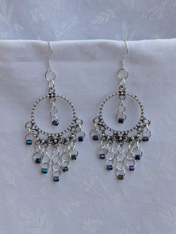 Moonlight Mirage Earrings: Pair of silver-colored hoop earrings with dangling silver jump rings and blue iridescent cylindrical beads.