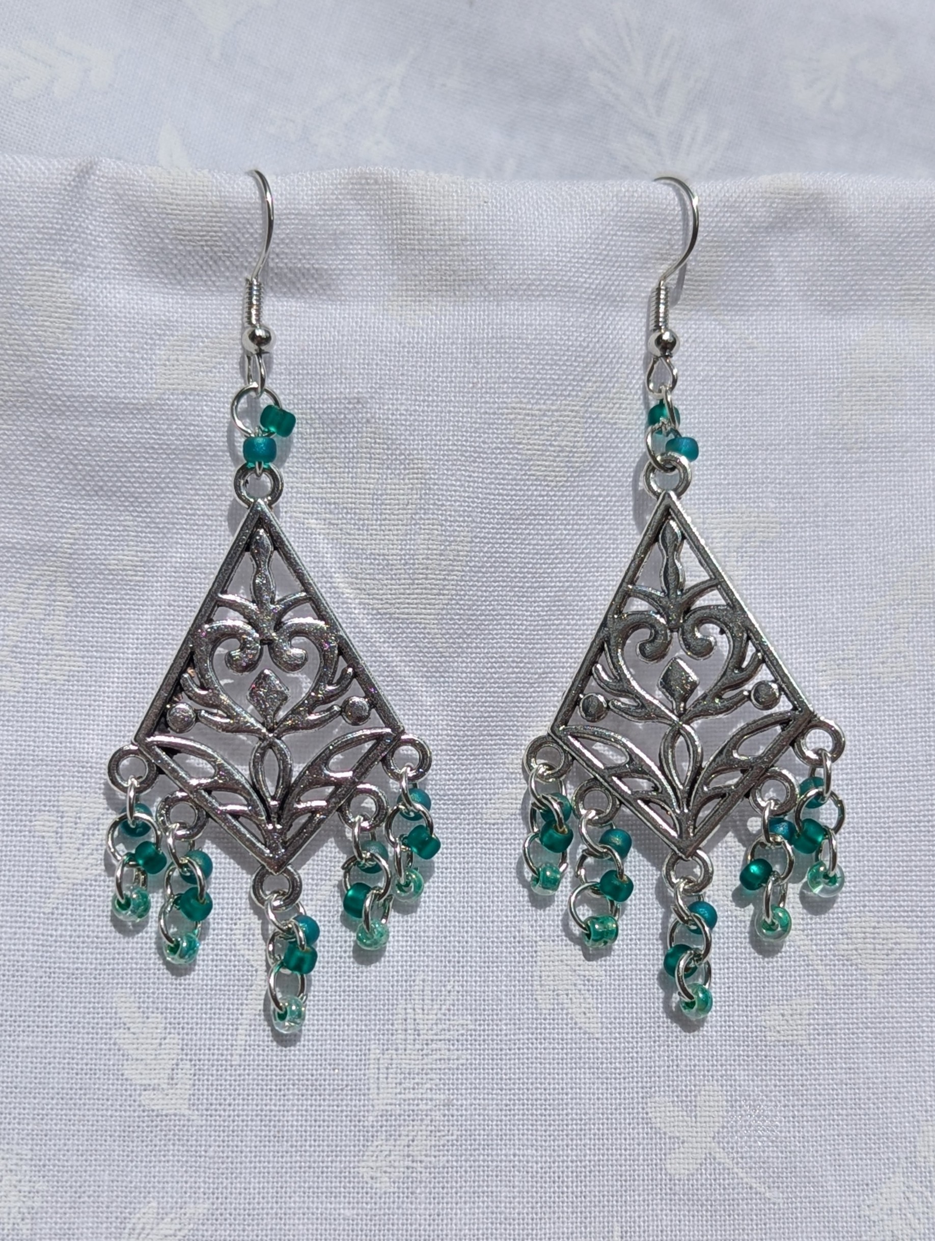Tideweaver earrings: Silver diamond-shaped filigree earrings with 15 cascading teal beads in frosted, matte, and translucent finishes, plus 2 teal accent beads above each ornament.