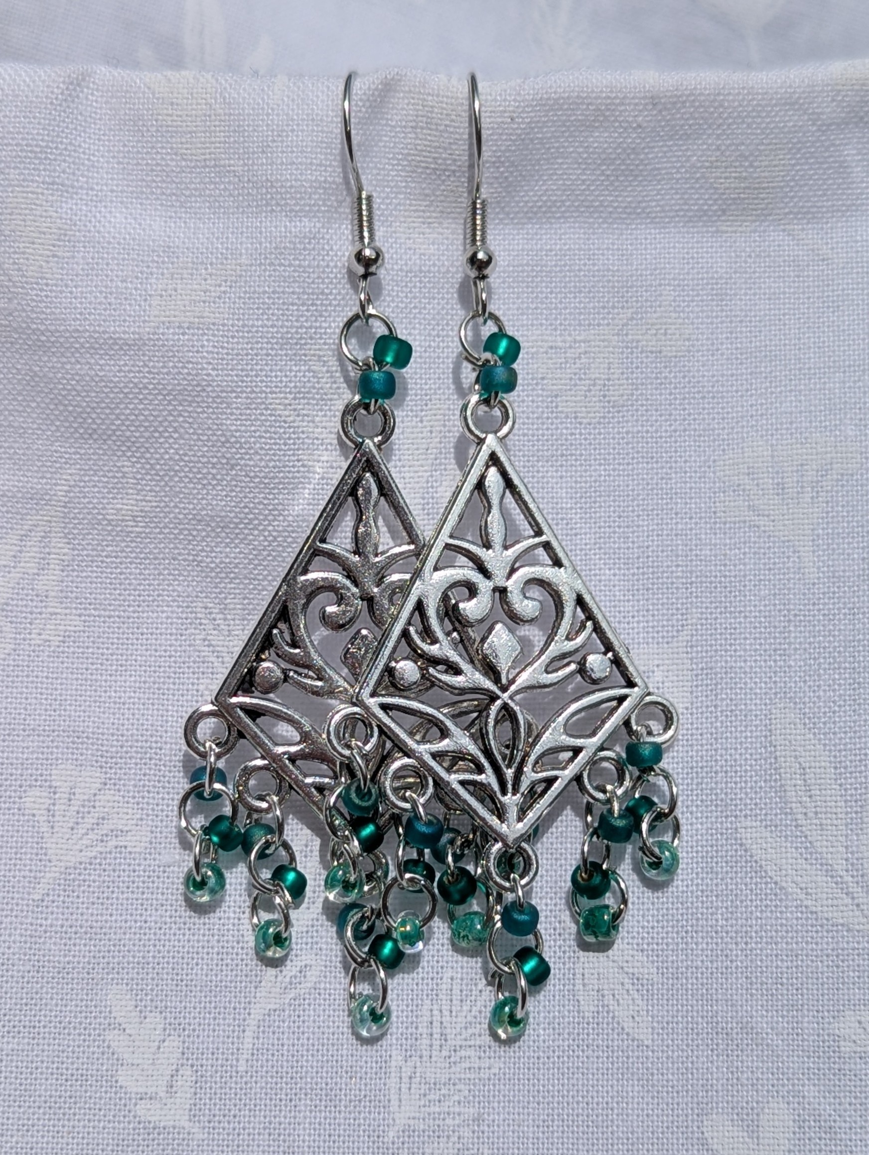Tideweaver earrings: Silver diamond-shaped filigree earrings with 15 cascading teal beads in frosted, matte, and translucent finishes, plus 2 teal accent beads above each ornament.