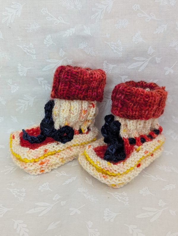 Handknit newborn baby booties in cream, red, and yellow with navy ties and a fiery red cuff, made from soft merino wool.
