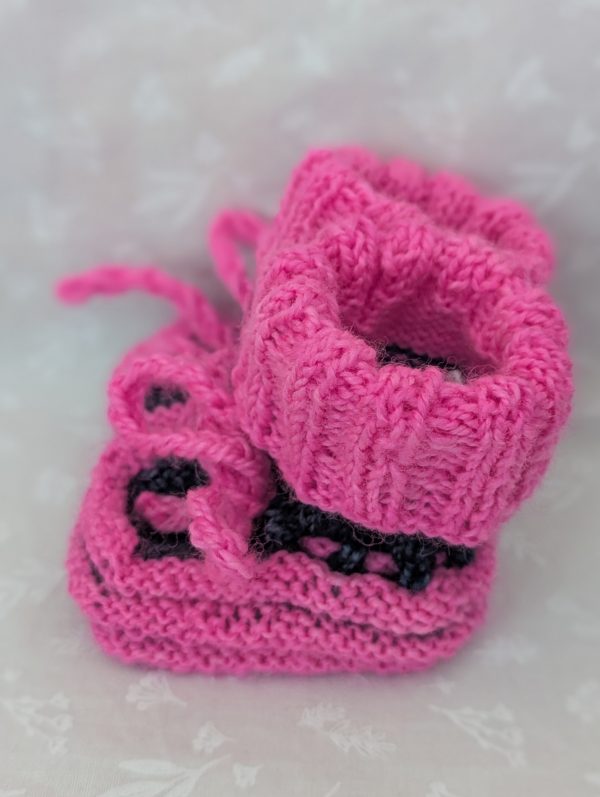 Pink handknit baby booties with blue/black accents, ribbed cuffs, and tied laces, made from soft merino wool for newborns.
