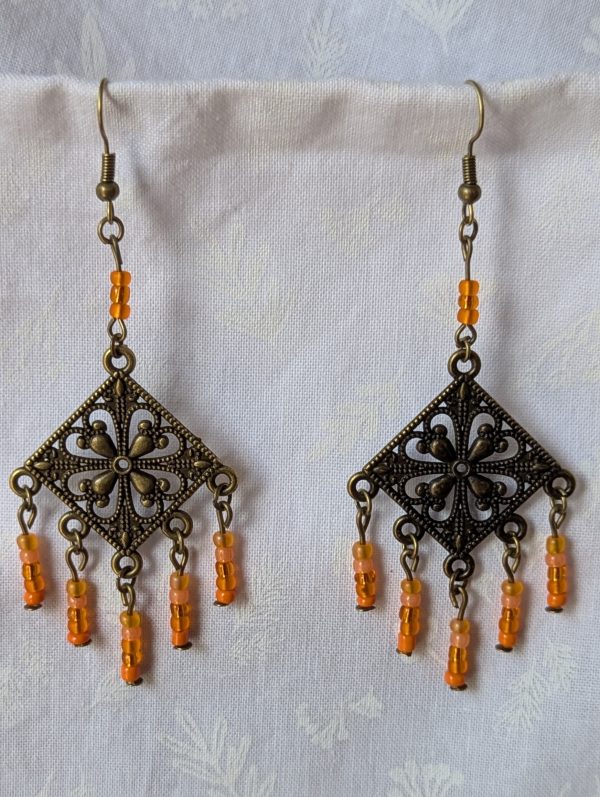 Orange beaded earrings, handcrafted with antiqued bronze square medallions and dangling orange glass beads in five textures, including translucent, frosted, and satin finishes. Hung on bronze ear hooks.
