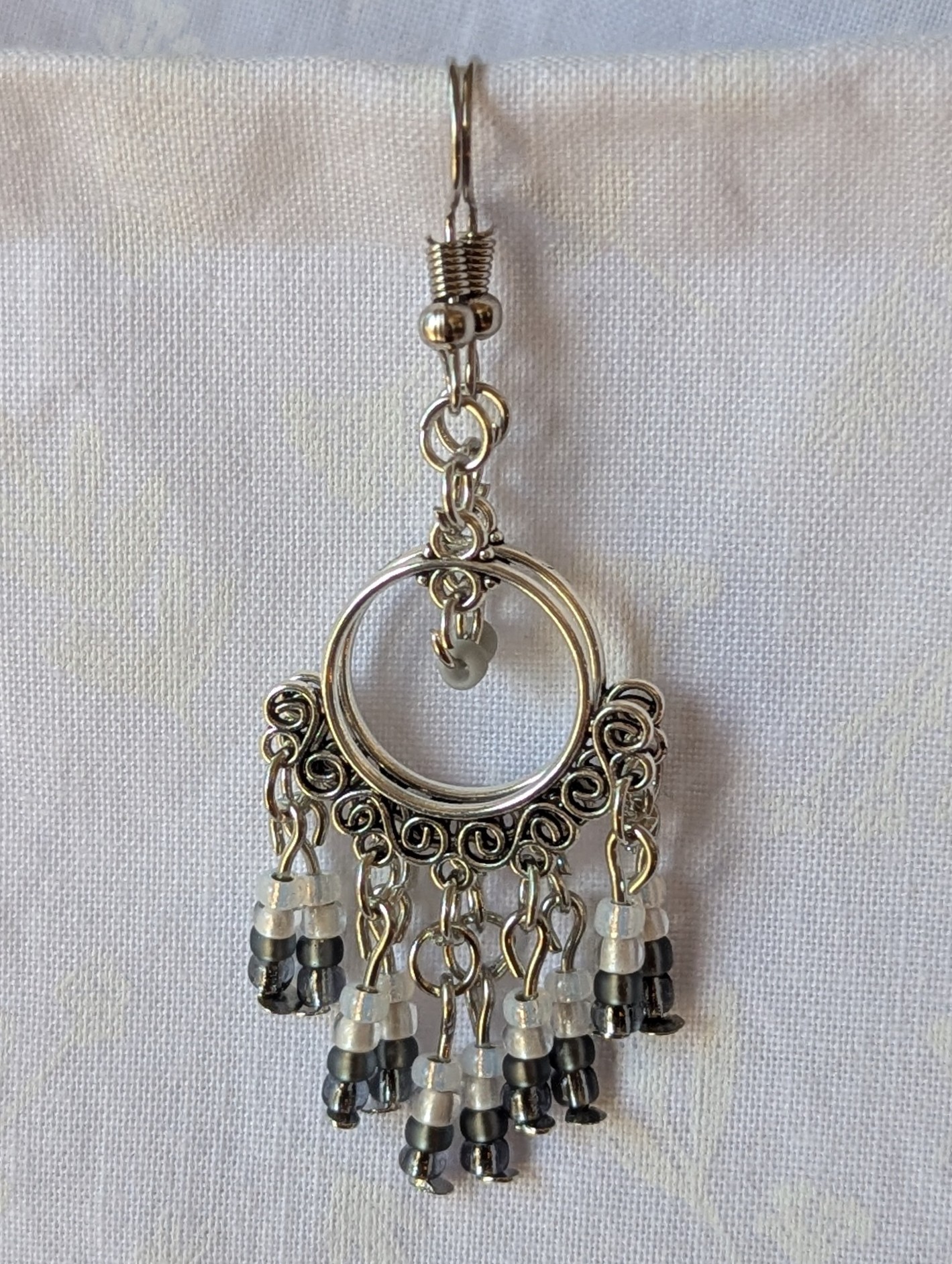 Pair of black and white earrings, handcrafted with silver crescent filigree hoops and dangling glass beads in a white-to-black gradient, including opal, frosted, matte, smoky, and black finishes. Hung on silver hooks.