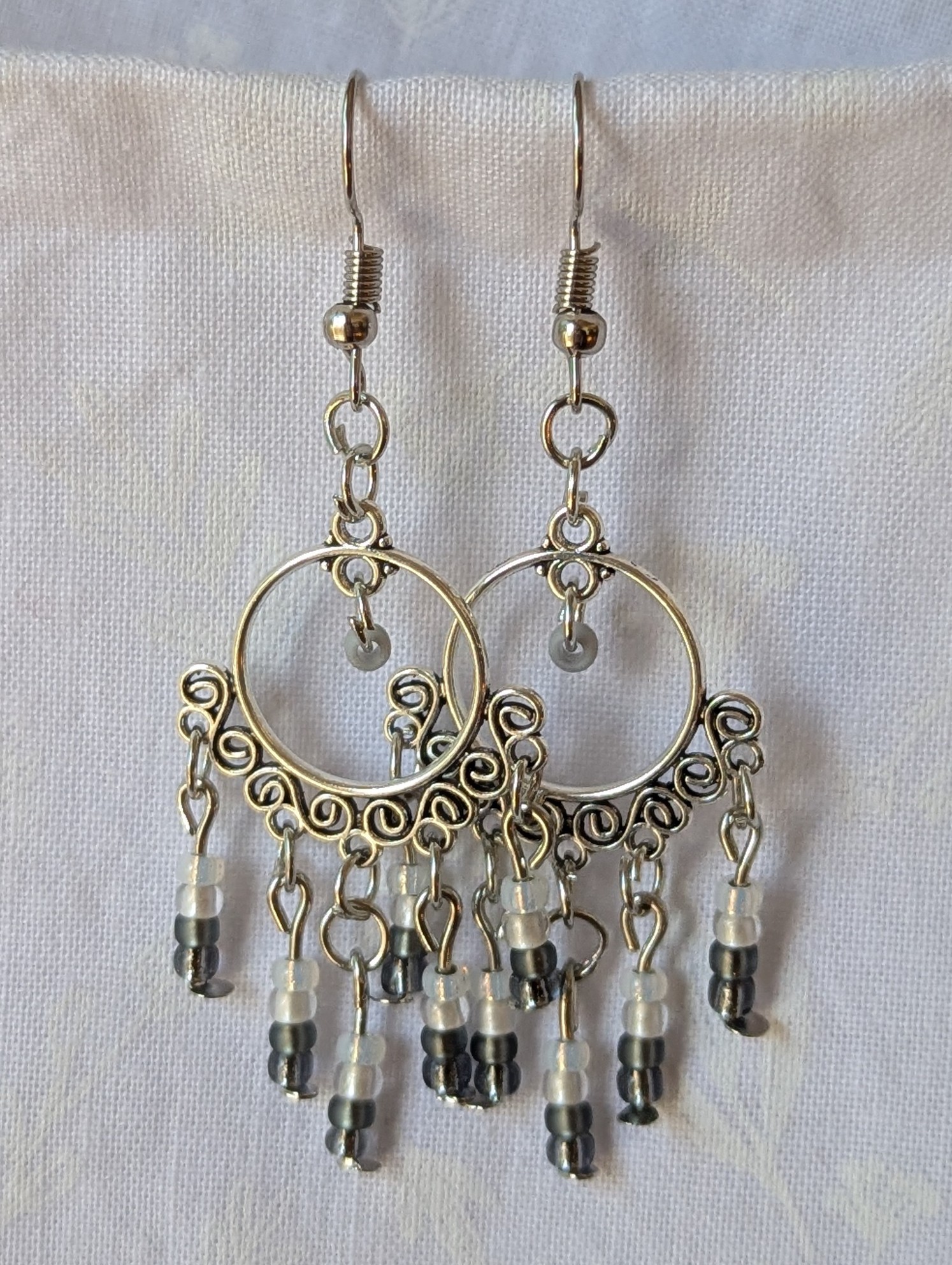 Pair of black and white earrings, handcrafted with silver crescent filigree hoops and dangling glass beads in a white-to-black gradient, including opal, frosted, matte, smoky, and black finishes. Hung on silver hooks.