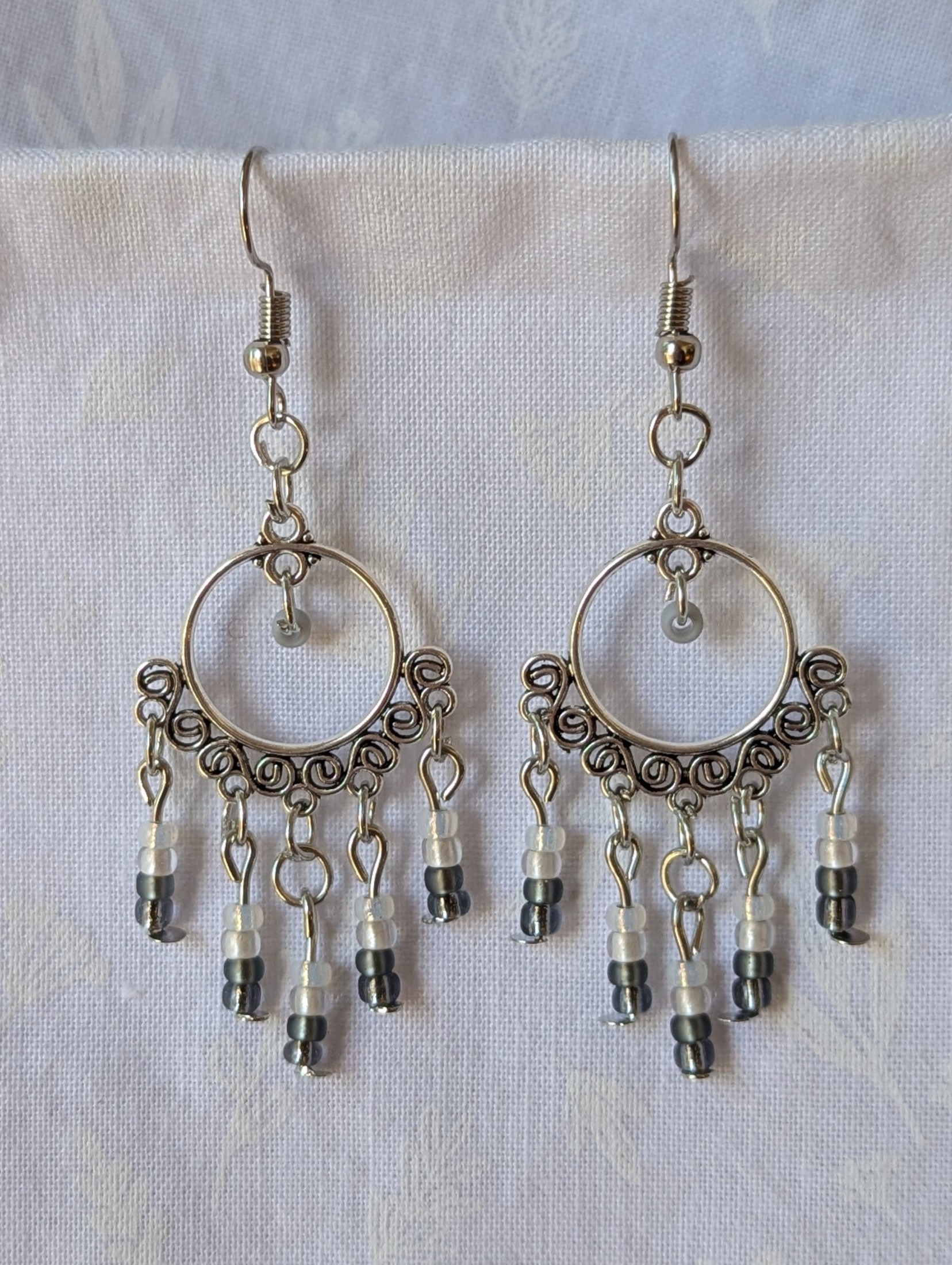 Pair of black and white earrings, handcrafted with silver crescent filigree hoops and dangling glass beads in a white-to-black gradient, including opal, frosted, matte, smoky, and black finishes. Hung on silver hooks.