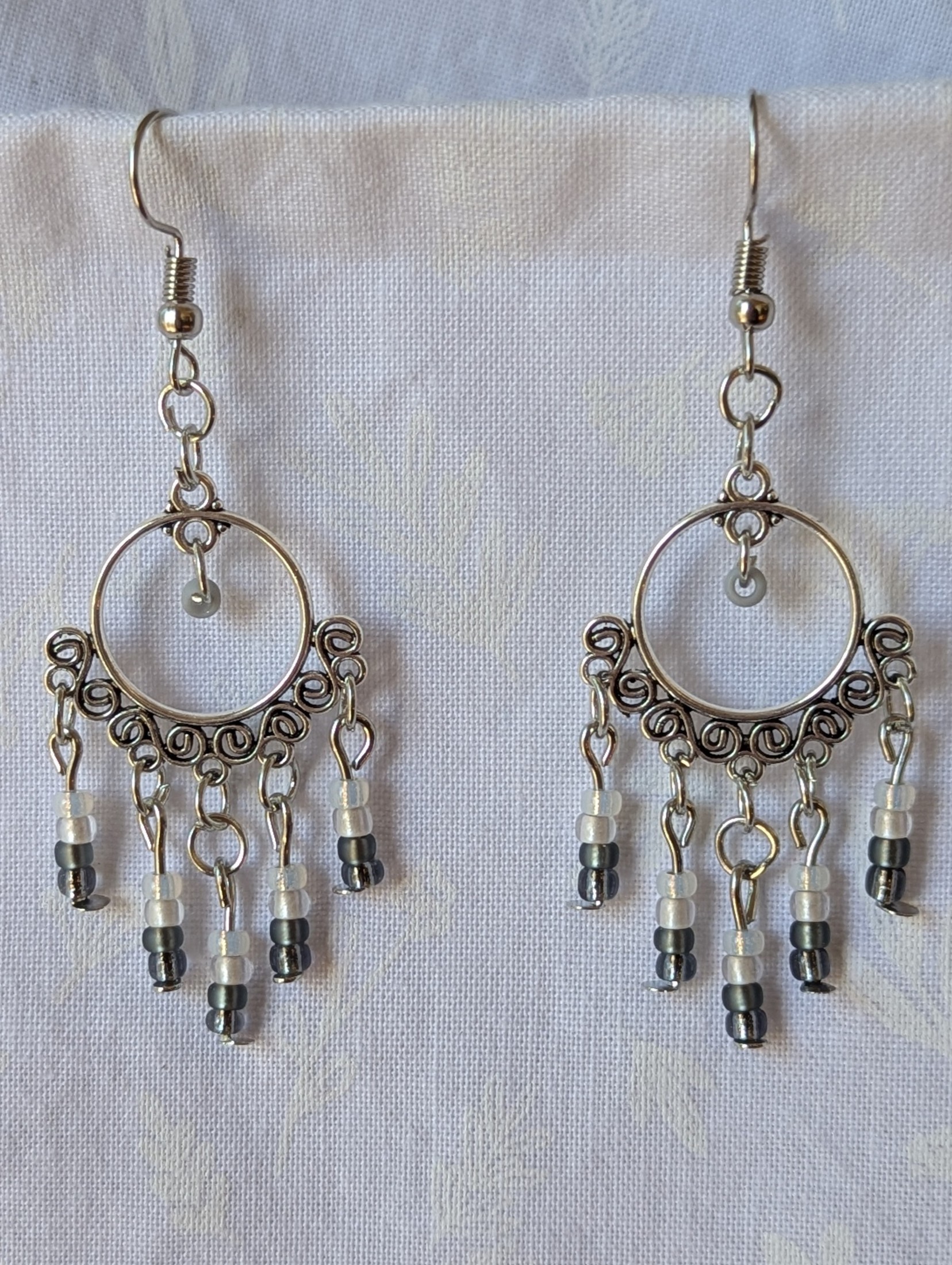 Pair of black and white earrings, handcrafted with silver crescent filigree hoops and dangling glass beads in a white-to-black gradient, including opal, frosted, matte, smoky, and black finishes. Hung on silver hooks.