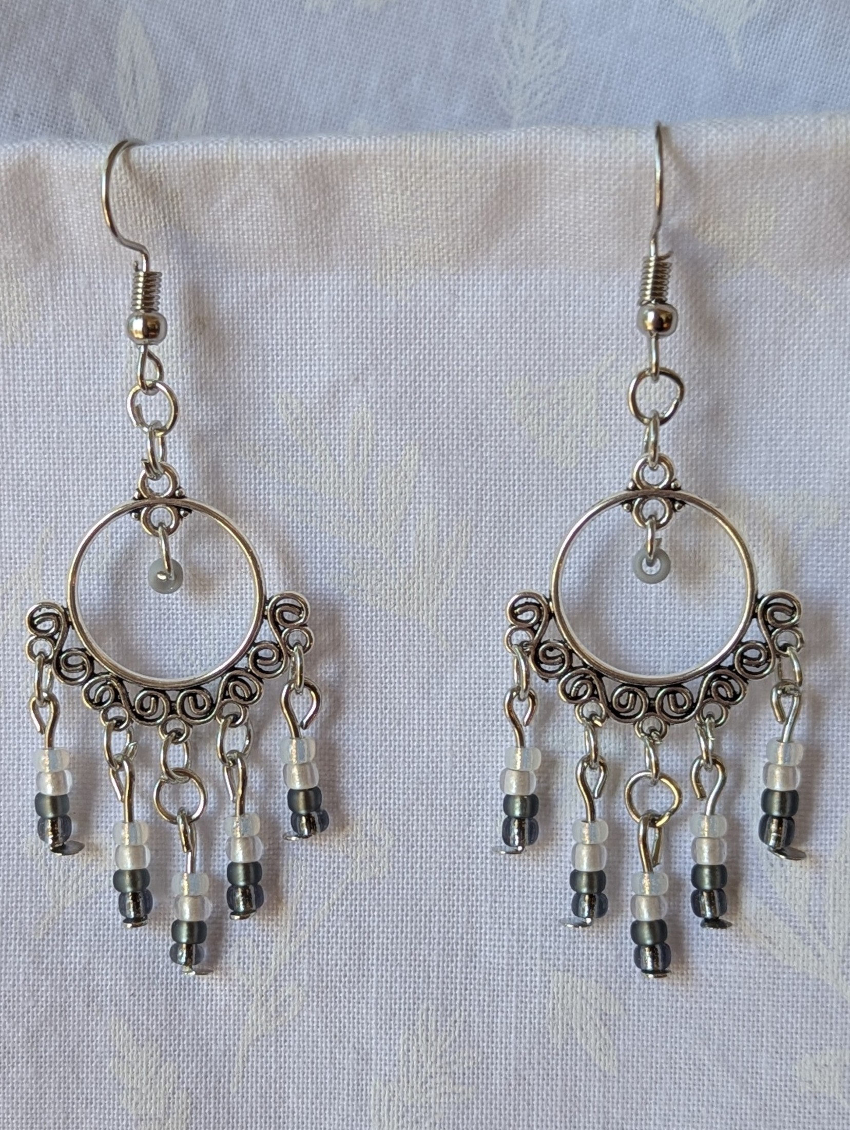 Pair of black and white earrings, handcrafted with silver crescent filigree hoops and dangling glass beads in a white-to-black gradient, including opal, frosted, matte, smoky, and black finishes. Hung on silver hooks.